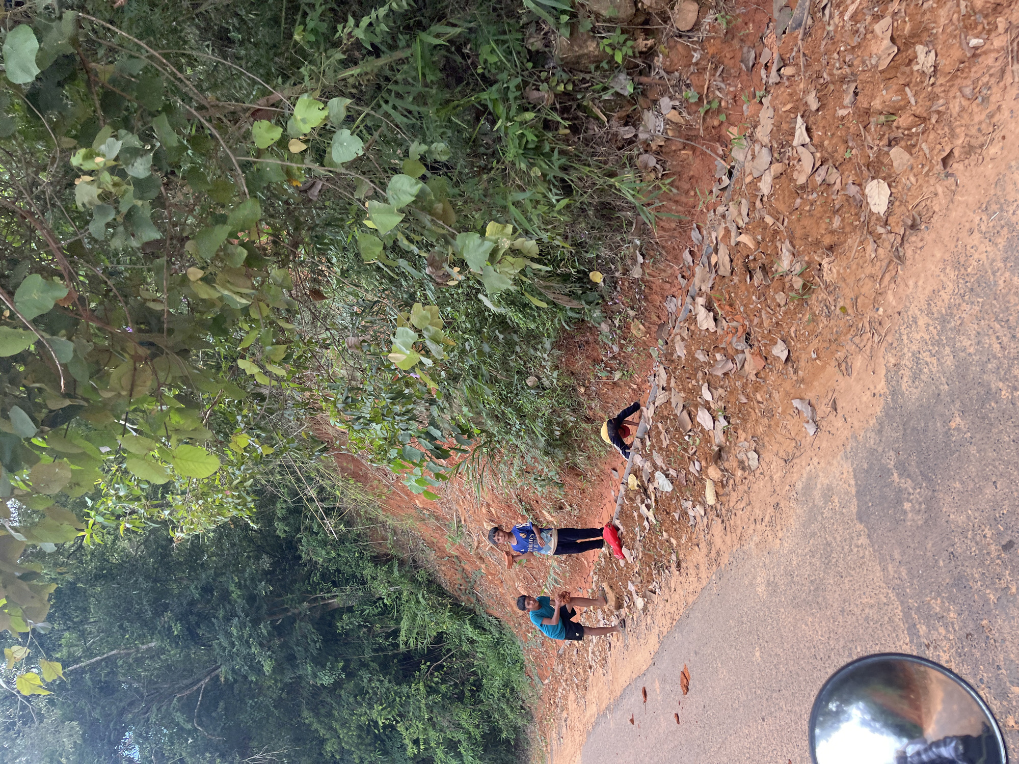 This is a medium shot, eye-level, outdoor photograph taken on a sunny day in Son Dien, Vietnam. The image captures a roadside scene with three children and a motorcycle mirror in the foreground.
On the left side of the paved road, a young boy wearing a blue t-shirt and black shorts is squatting down. To his right, a slightly older boy in a blue and white jersey and dark pants stands looking towards the camera. Further to the right, a third child, wearing a yellow shirt and red head covering, is partially visible, hunched over near the edge of the road, possibly interacting with something on the ground.
The roadside is composed of red dirt, scattered with dry leaves and some broken pieces of brick. Dense green foliage and trees line the embankment behind the children, creating a natural backdrop. A portion of a motorcycle's circular rearview mirror is visible in the bottom left corner of the frame, reflecting a blurred image of what appears to be the photographer. The overall scene suggests a rural or semi-rural setting.