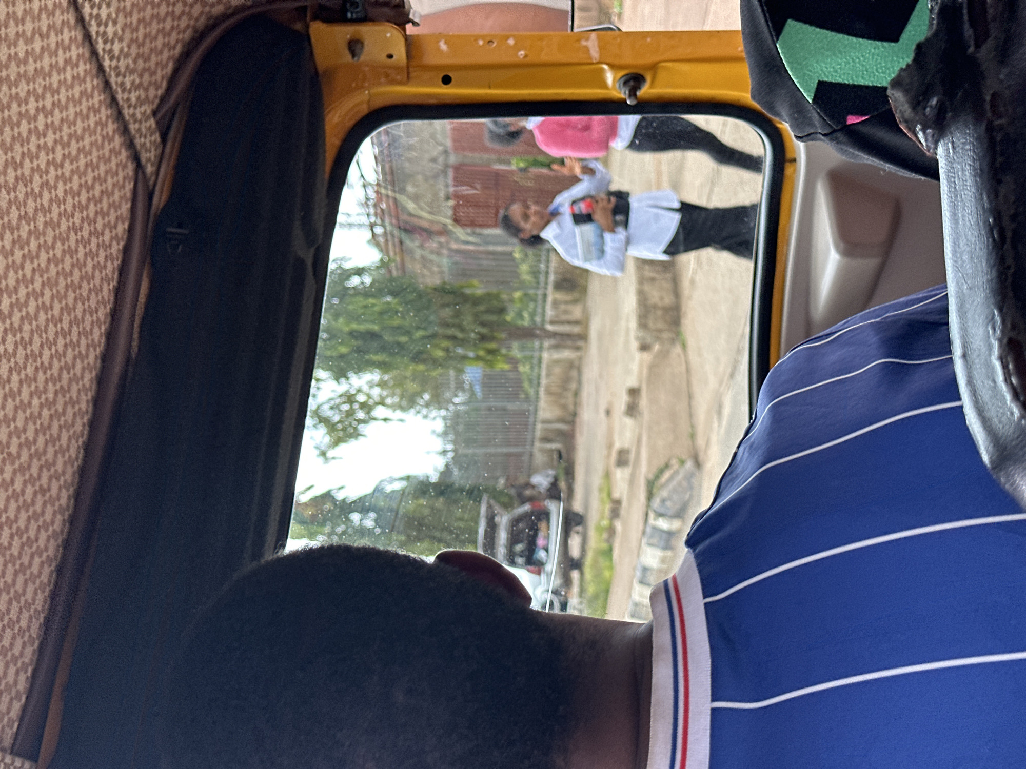 The image depicts a scene from within an auto-rickshaw, looking out of the side window. The primary subjects are two individuals visible through the window. One person, wearing a pink shirt and black pants, is blurred but seems to be gesturing. Another individual, in the foreground through the window, is wearing a white shirt and dark pants and appears to be holding a book or tablet and a phone. They are looking down and appear to be walking or standing on a street.
The setting is an outdoor environment, likely a street scene in Nigeria, given the location context of Bar Jirgi Summa. We can see buildings with red tiled roofs and a paved street. Trees and foliage are visible in the background, suggesting a daytime scene, possibly late morning or afternoon given the lighting. The auto-rickshaw's interior is partially visible, with textured brown upholstery and yellow paint on the exterior frame. A person's head and shoulder, wearing a blue and white striped shirt, are in the foreground, obscuring part of the view from inside the vehicle. The overall mood appears to be one of transit, with a focus on the external world observed through the window. There is no visible text in the image.