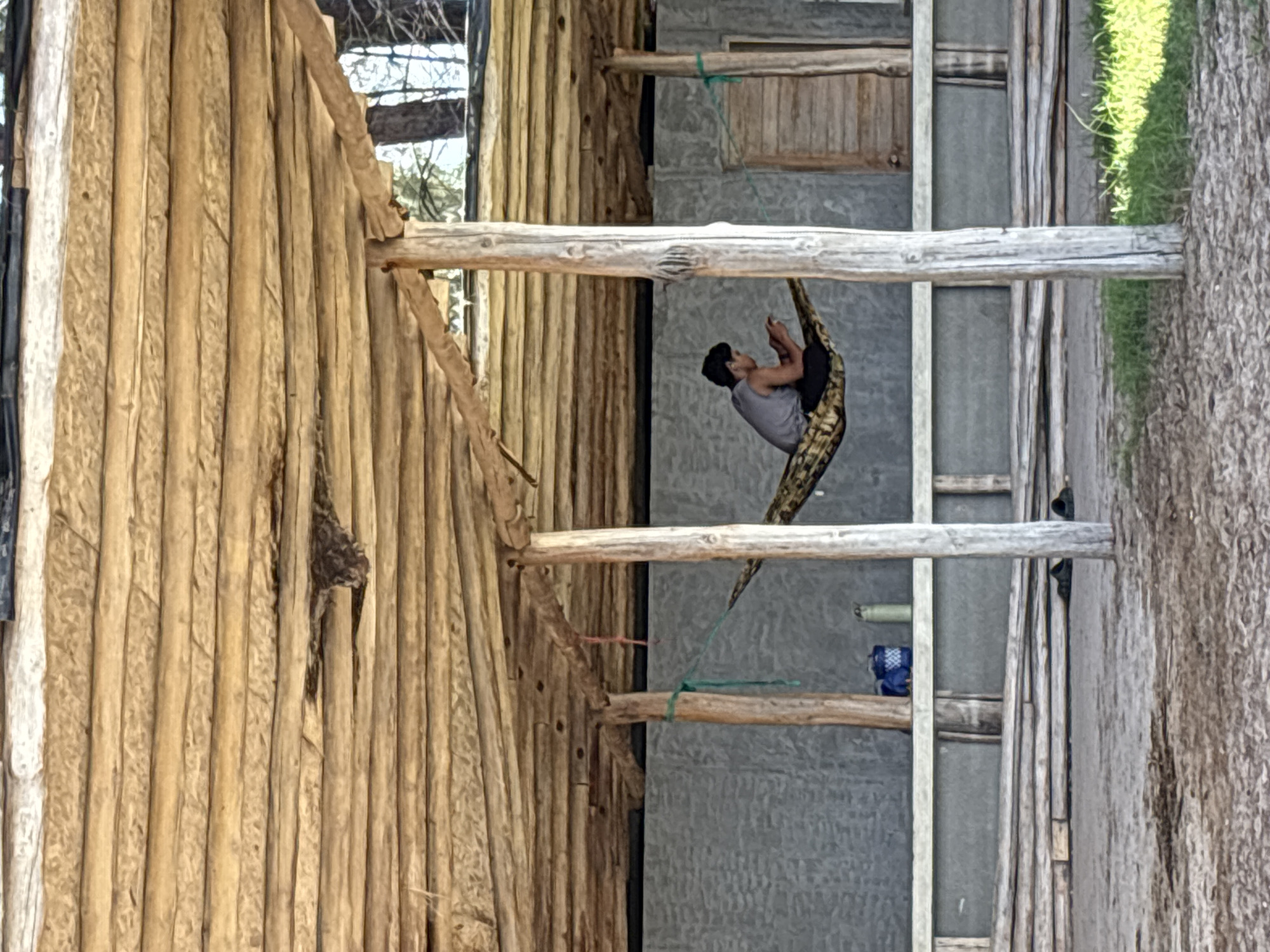 A man is suspended in a yoga hammock apparatus inside an outdoor structure. The structure is made of raw lumber and rough-hewn wood. The location is near Choeng Thale, Thailand. The yoga hammock apparatus hangs between two horizontal lumber beams. The man faces left, with his legs bent up. He is wearing a gray shirt and dark shorts. A tree trunk is visible to the right of the structure.