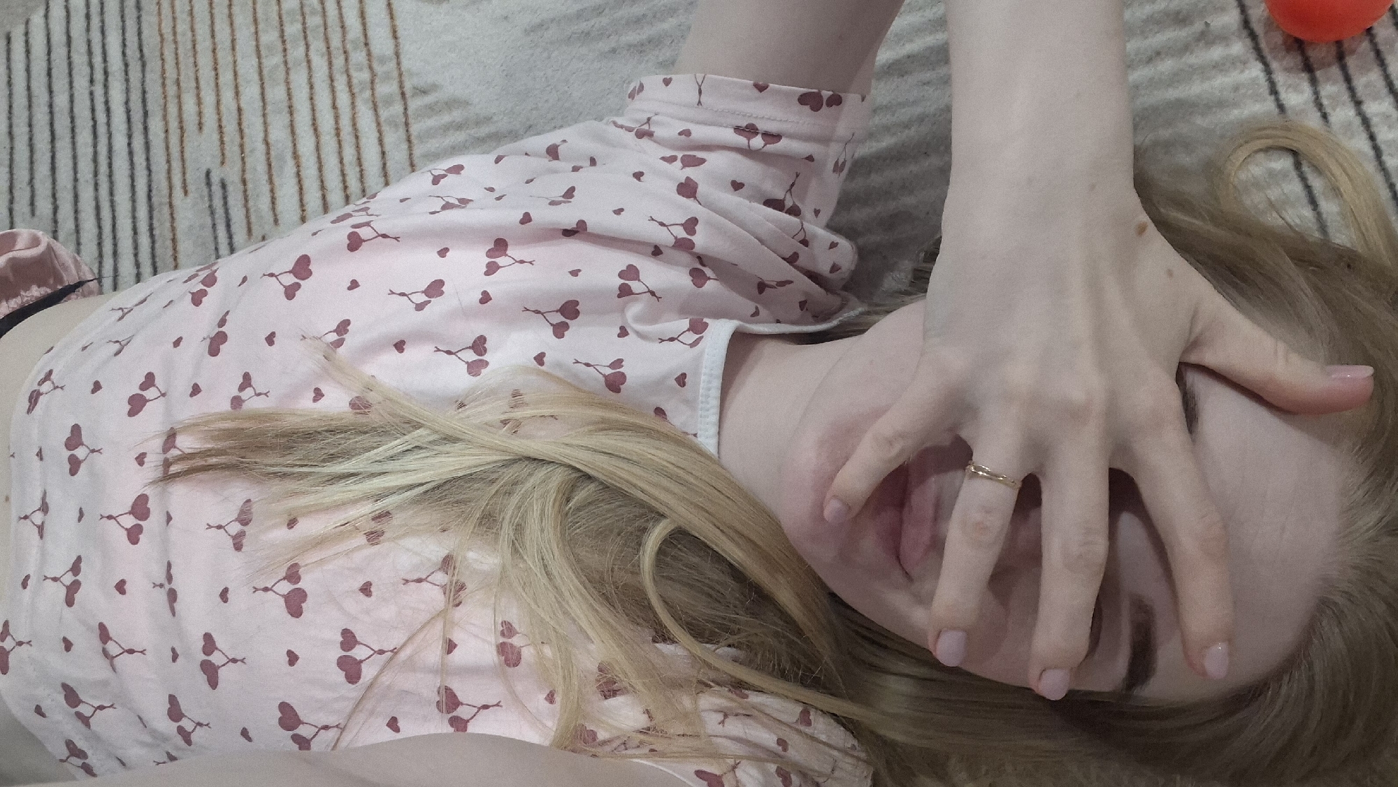 A fair-skinned person with blonde hair covers their face with their hand, revealing their lips, while lying on a striped surface. The person is wearing pink pajamas patterned with cherries. The person appears to be wearing a gold ring on their ring finger. An orange object is present in the upper right corner of the image. The location is Chaikovsky, Russia.