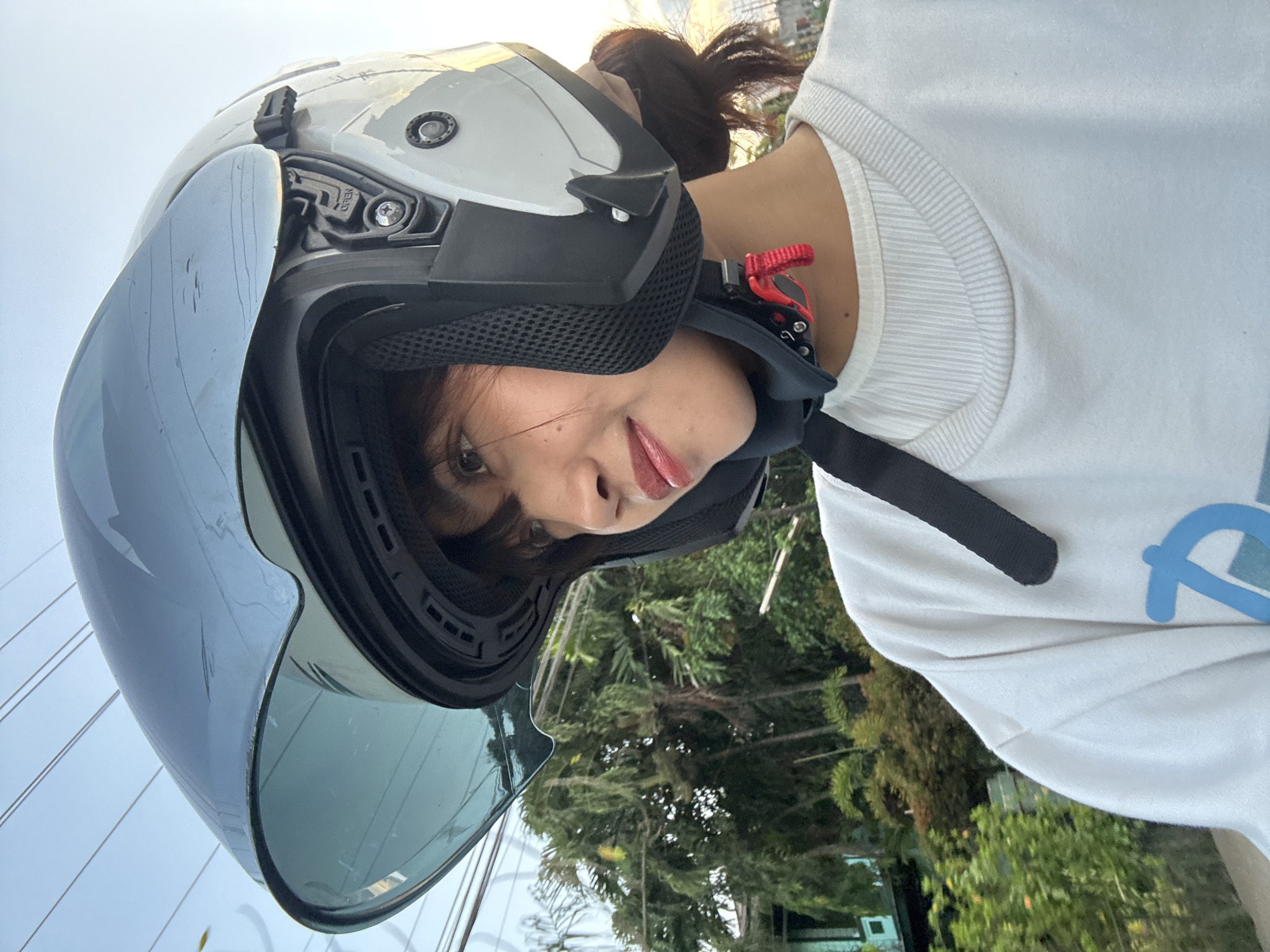 A young woman wearing a white long-sleeved shirt and a white helmet with a tinted visor is pictured in what appears to be a residential area in Davao City, Philippines. She is looking off to the left, with a slight smile on her face. The background shows lush green trees and foliage, with some houses visible in the distance. Power lines are also visible against the sky, suggesting an urban or semi-urban setting. The lighting indicates it could be late afternoon or early evening. The woman is likely riding a motorcycle, given the helmet.