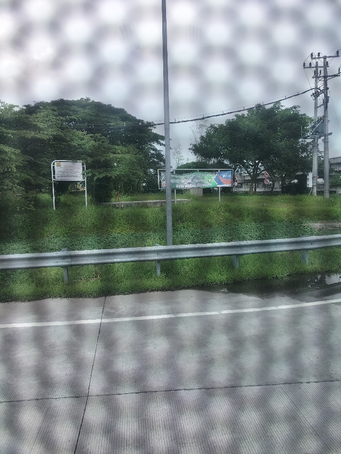 The image shows a view of a road in Kebomas, Indonesia (latitude: -7.168, longitude: 112.596) from a car. There is a gray metal guardrail along the edge of the road, and green grass is growing on the embankment. A single white line is painted across the gray asphalt road. A large green tree is visible in the background, and a sign with Indonesian text is partially visible next to a tall metal pole with wires running to it. The sky is overcast.