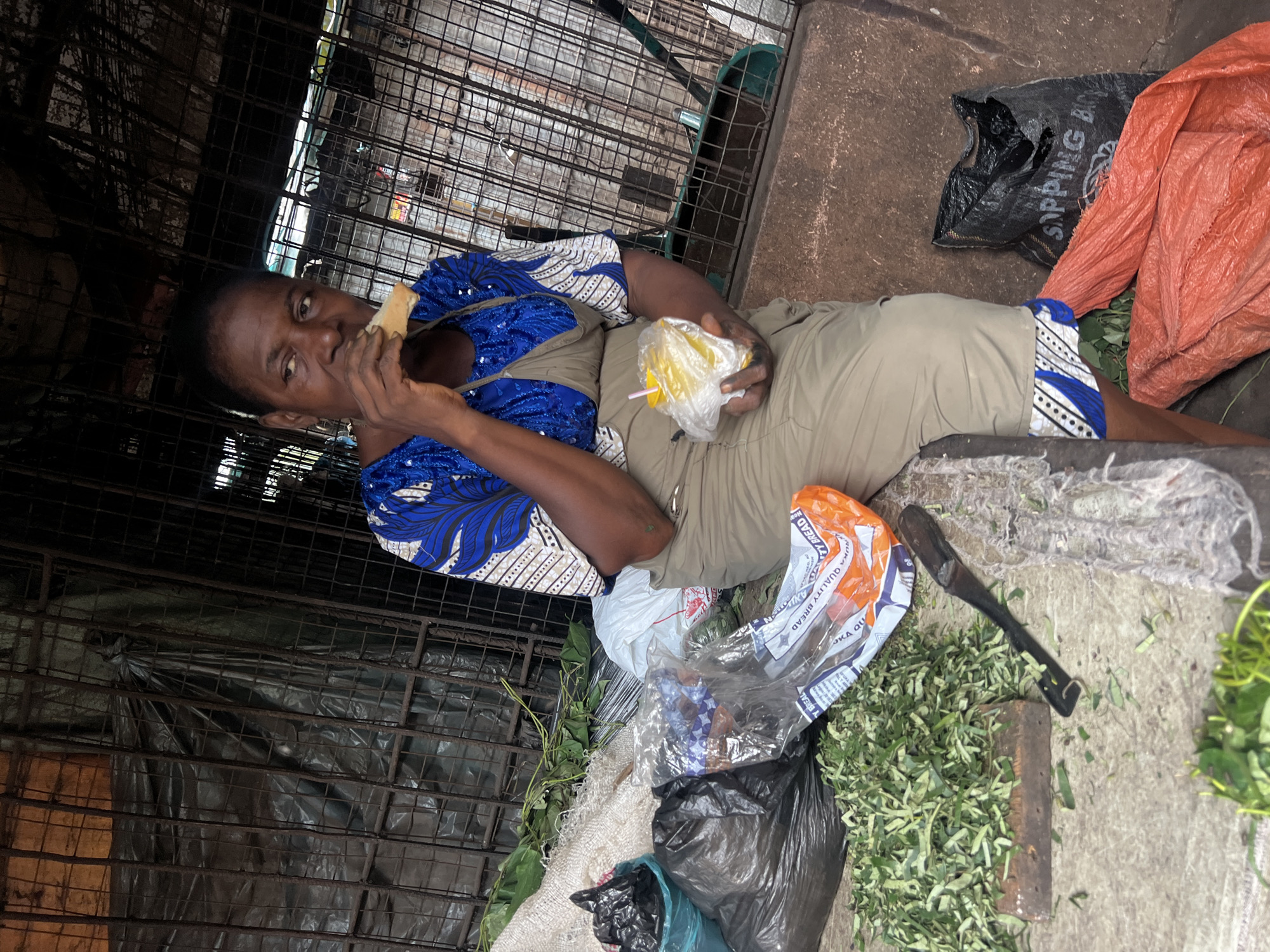 A woman sits on the ground in Nkpor, Nigeria. She wears a blue and white patterned top and khaki pants with a blue and white patterned hem. She eats a piece of bread and holds a plastic bag containing yellow food items. Next to her are bags of goods, green leafy vegetables, and a tool with a black handle. The background consists of a metal cage and a concrete surface.