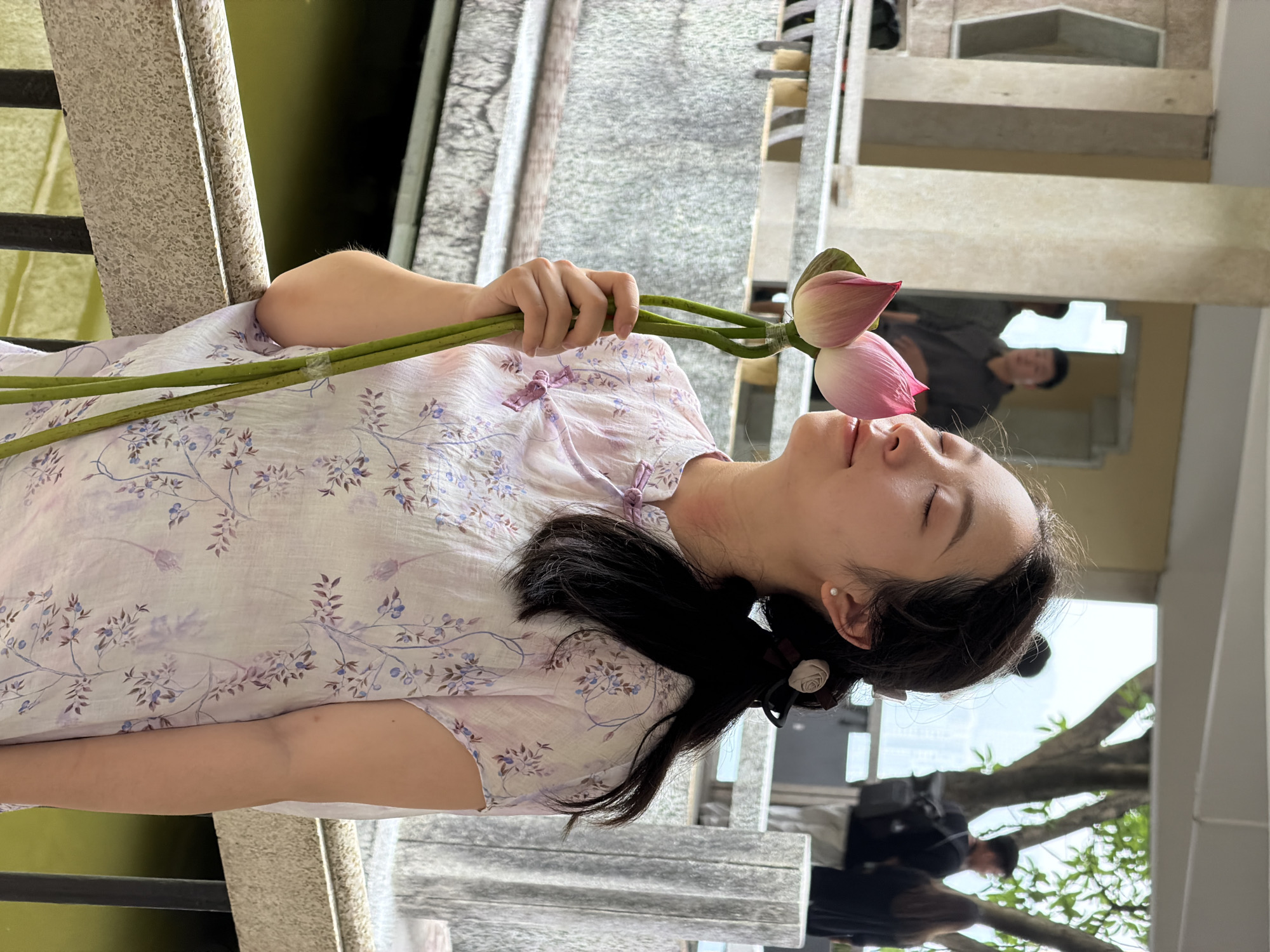 A young Asian woman with dark hair tied back in a ponytail is standing outdoors, holding two lotus flowers up to her face and smelling them with her eyes closed. She is wearing a light pink dress with a floral pattern and has a small white earring in her visible ear. The background shows a body of water, a stone railing, pillars, and several blurred figures of people walking in the distance. The location is Yuzhong District, China.