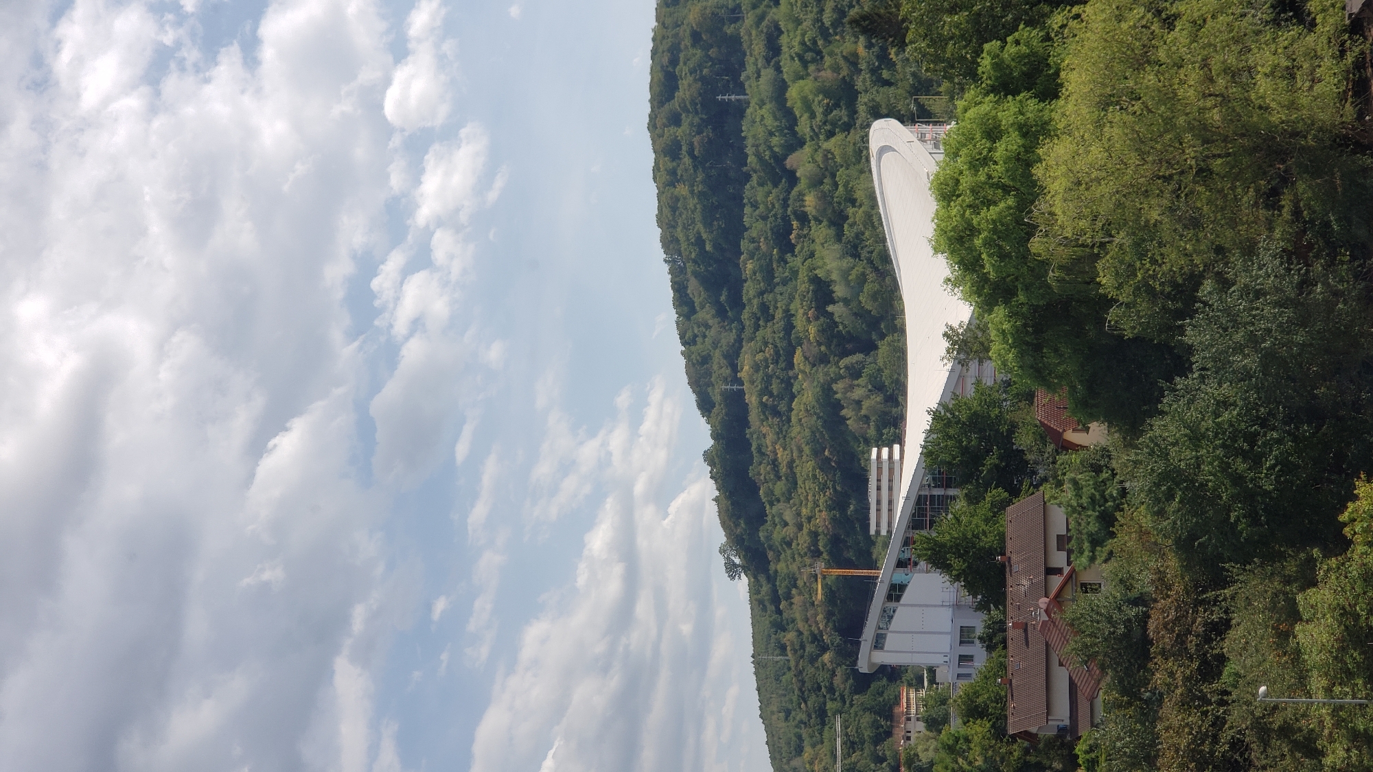 The image depicts a modern, curvilinear building with a white, undulating roof, possibly a stadium or arena, under construction. The building is situated on a slope amidst a densely wooded hillside. Several residential buildings with red tile roofs are nestled in the foreground, partially obscured by lush green trees. A yellow construction crane is visible behind the main structure. In the distance, the hillside is covered with a vast expanse of green trees, with a few power line pylons visible through the foliage. The sky is partly cloudy with soft, diffused sunlight. The scene is likely an aerial or elevated view of the area, possibly showing the stadium complex in Prešov, Slovakia.