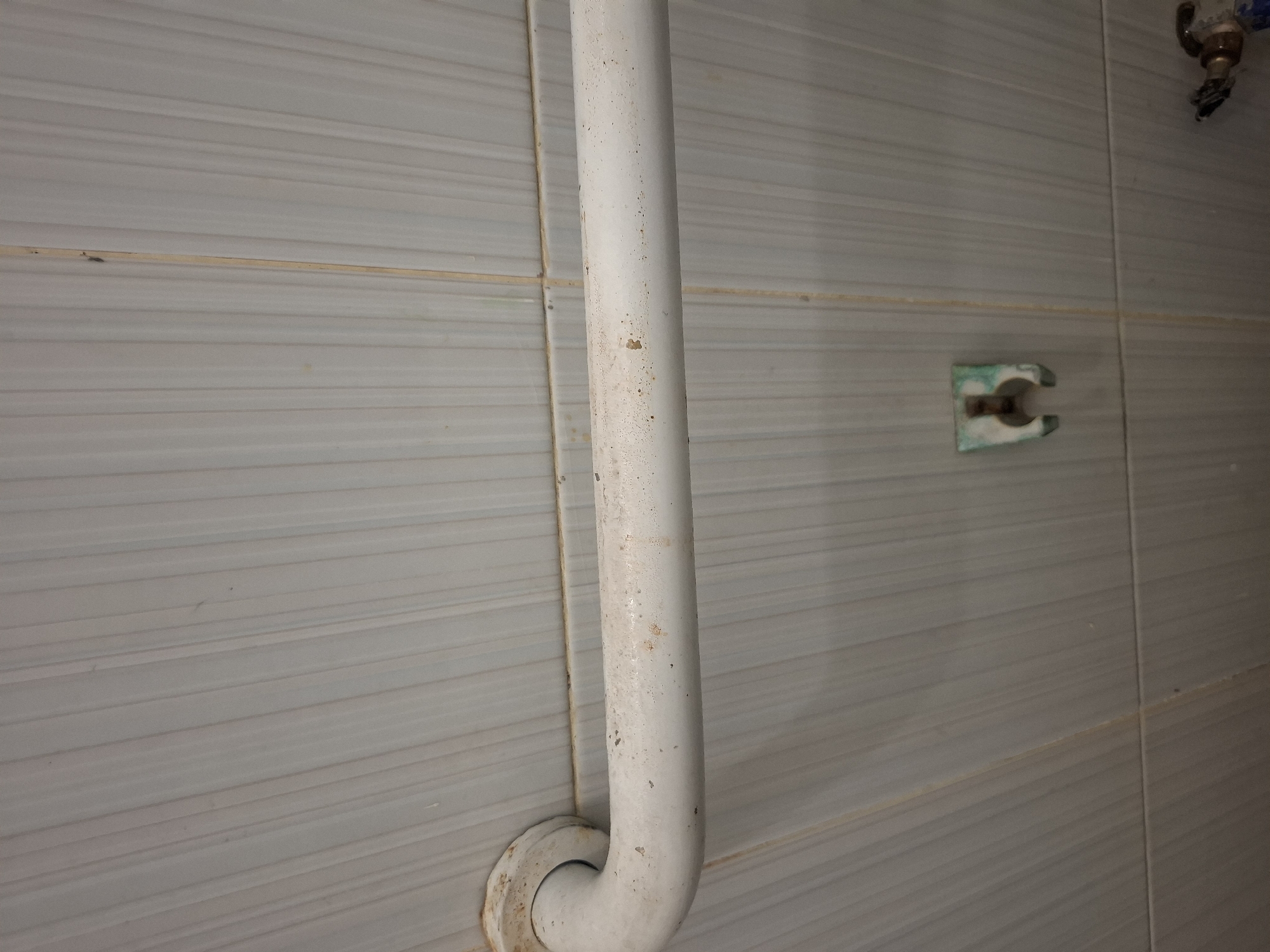 A close-up, head-on shot captures a white grab bar mounted horizontally on a tiled wall in Meureudu, Indonesia. The grab bar appears to be made of metal and shows signs of wear, with some discoloration and speckling along its surface. The tiles are light gray and arranged vertically, with thin grout lines. To the right of the grab bar, a small, green bracket is attached to the wall. The lighting is even, indicating an indoor setting, likely a bathroom or shower area. The scene is static and devoid of human presence.
