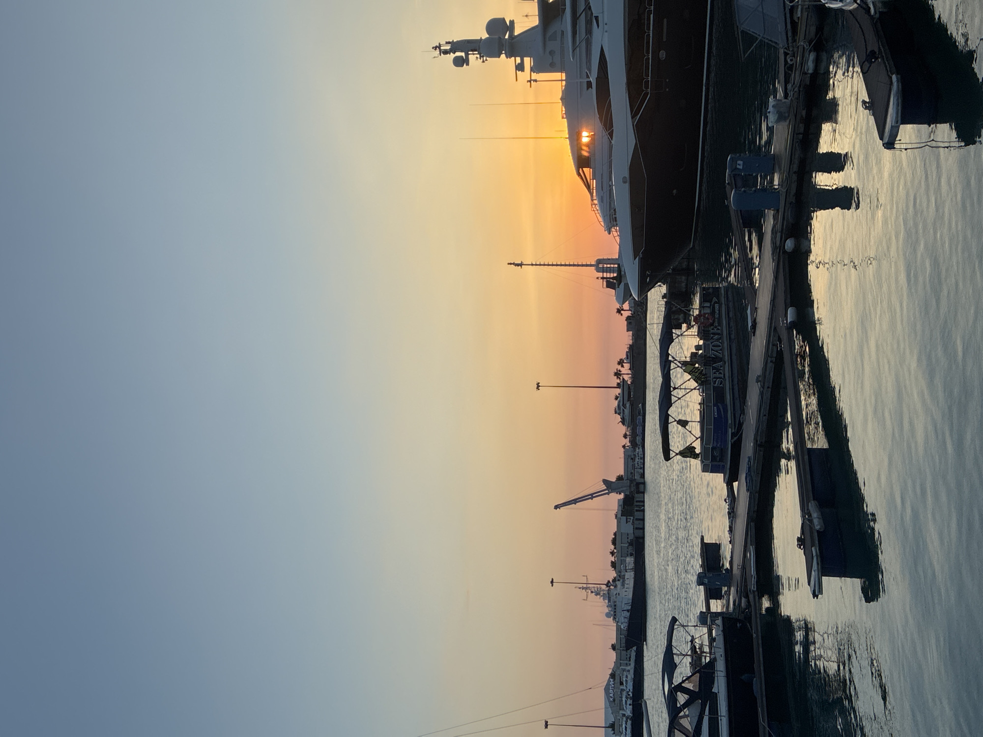 This is a vertical, eye-level, medium shot of a harbor in Sochi, Russia, at sunset. The sky transitions from a pale blue at the top to a vibrant orange and pink as it nears the horizon. The water in the foreground is calm, reflecting the colors of the sky and the surrounding boats. Several yachts and smaller boats are docked in the harbor. The largest yacht, prominently featured on the right, is a luxury vessel with multiple decks and a dark hull. To the left, a crane stands tall, indicating ongoing port activity. In the distance, more boats, buildings, and palm trees are visible along the coastline. The overall scene is peaceful and serene, capturing the beauty of a sunset over the Black Sea.