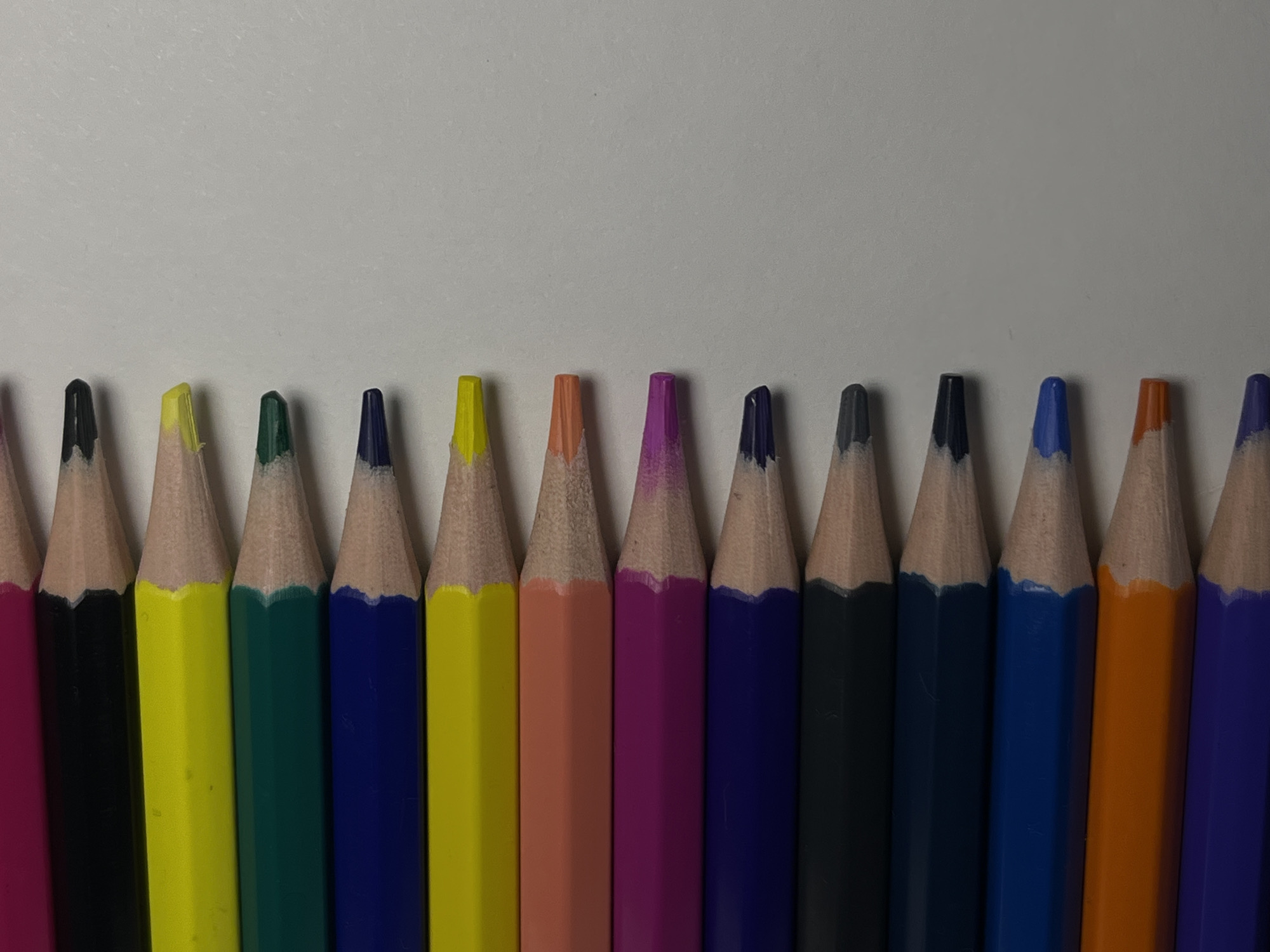 A collection of colored pencils is arranged in a row on a light surface. The colors are, from left to right: magenta, black, yellow, green, dark blue, yellow, orange, pink, dark blue, black, dark blue, light blue, orange, dark blue. The pencils are sharpened and positioned close together. The background is a plain, light-colored surface. There is no indication of location in Фоки, Russia or any people or actions visible in the image.