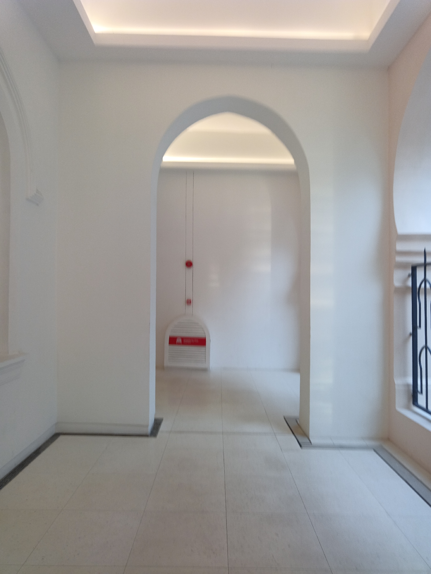 The image shows an indoor hallway with a tiled floor and white walls. An arched doorway leads into another section of the hallway, which is illuminated by recessed ceiling lights. A red fire alarm and a fire sprinkler system are mounted on the wall inside the arched doorway. A decorative grate is visible on the floor near the base of the wall on the right side of the image. The overall scene suggests a modern building interior. The location is identified as Kuala Lumpur, Malaysia, likely a public space within a building such as a hotel, mall, or office building. There are no people or animals present in the image.