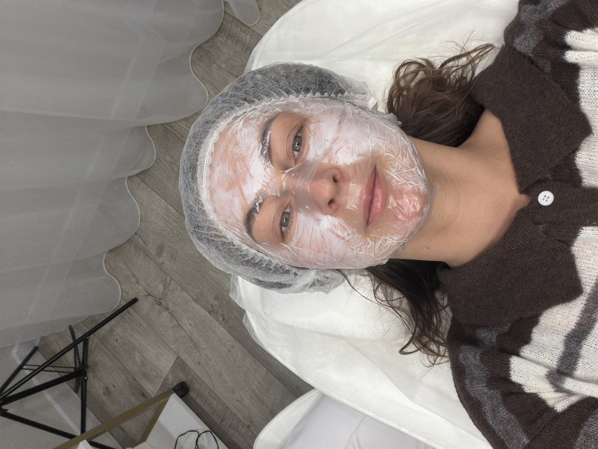 A woman with a white face mask and a hairnet lies on a white surface, possibly a treatment table, in a bright indoor setting. She is wearing a brown and white sweater. A light-colored wooden floor is visible with a tripod in the lower left corner and a sheer white curtain along the left side. The location is Chaikovsky, Russia.