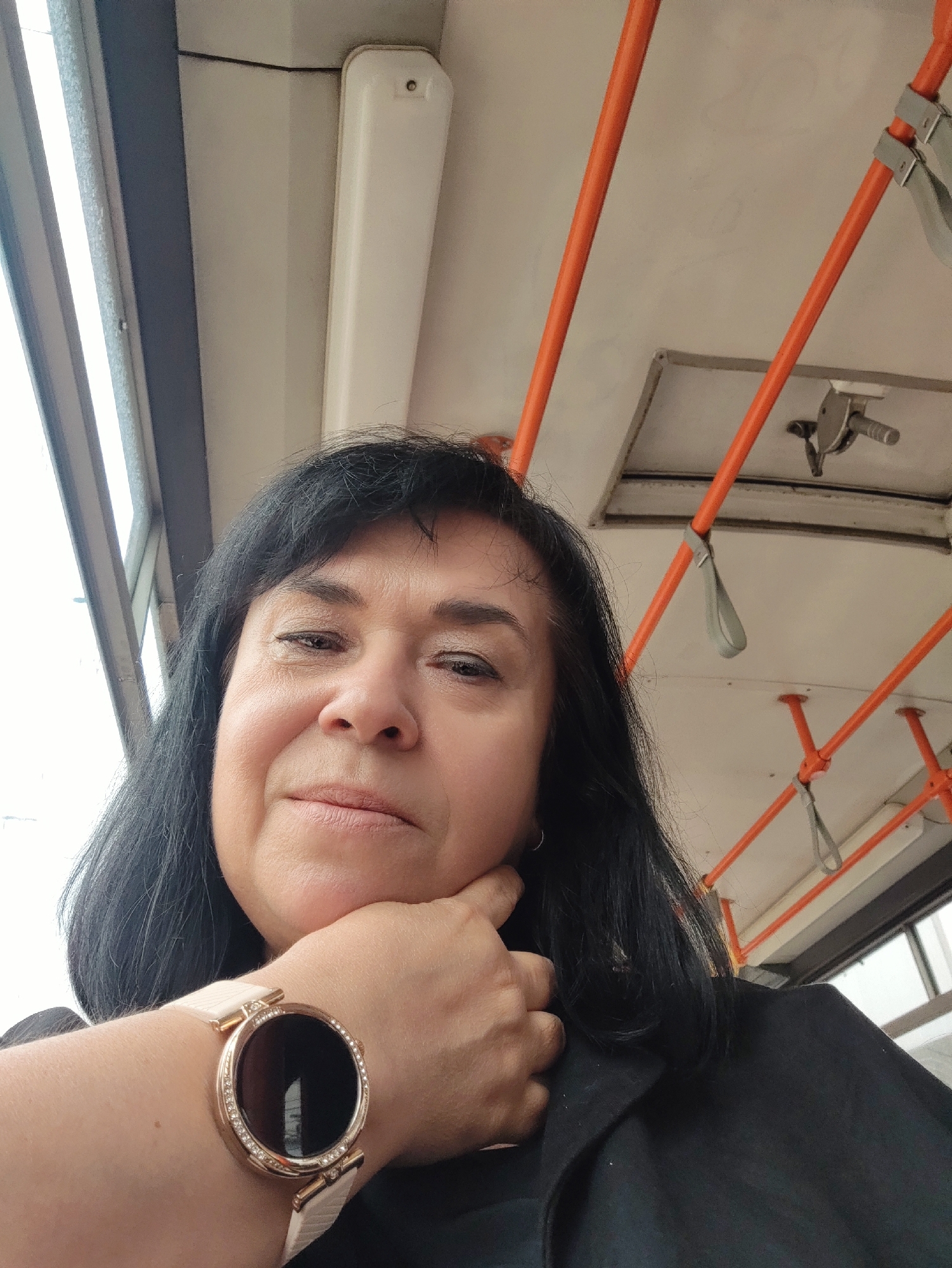 An outdoor, medium shot selfie shows a woman with dark hair and brown eyes resting her chin on her left hand, which is adorned with a rose gold smartwatch. She is wearing a black jacket. The background suggests she is inside a public transportation vehicle, possibly a bus or train, with orange grab bars and a white ceiling visible. The lighting is bright, indicating it is daytime. The specific location within the city of Unknown is not determinable from the image.