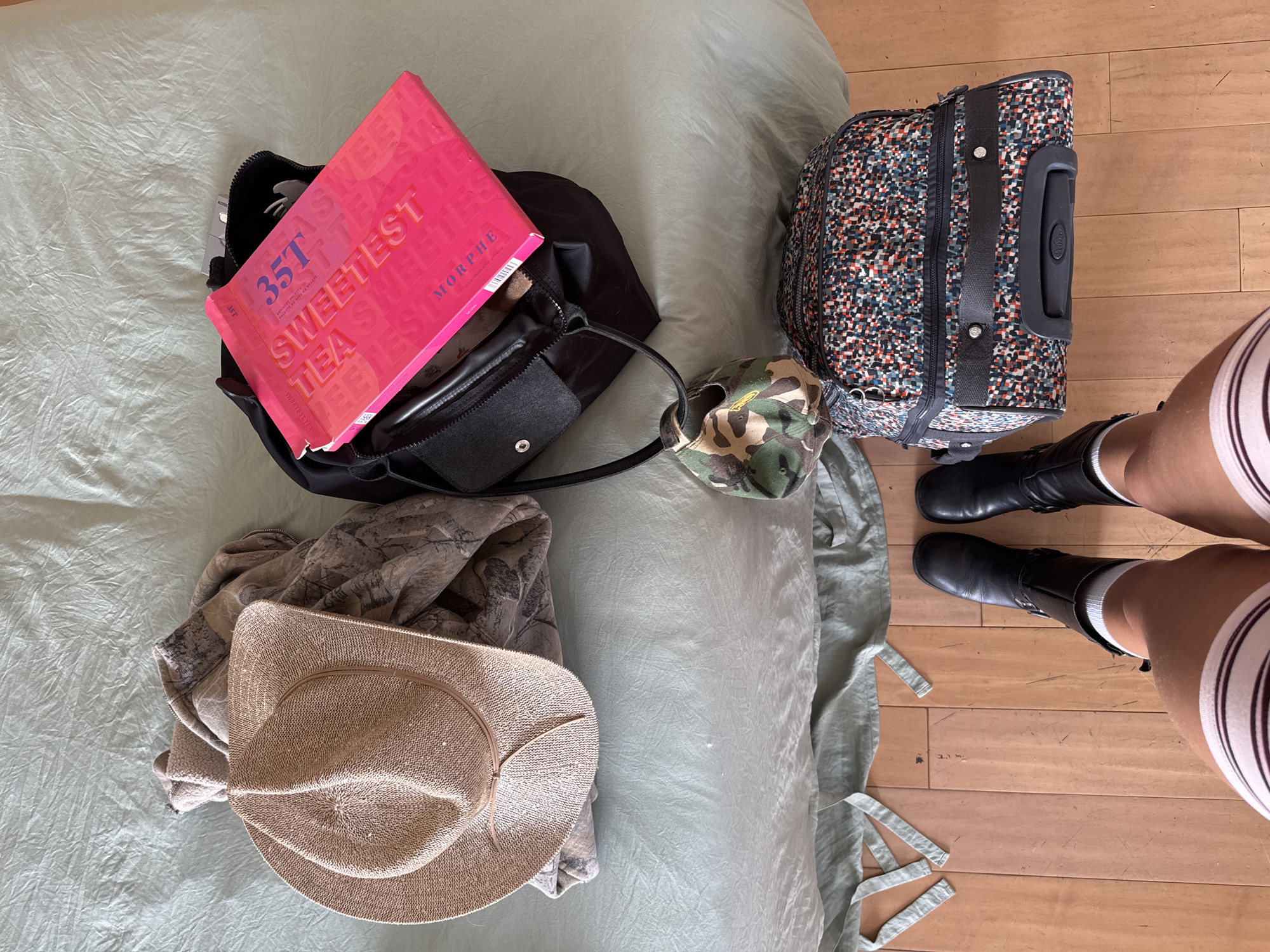 The image captures a scene on a bed in what appears to be a home or hotel room, in Los Angeles, United States of America. The bed is covered with a green sheet. Items arranged on the bed include a black handbag with a pink "35T Sweetest Tea" Morphe palette inside, a camouflage baseball cap, a patterned suitcase, a tan cowboy hat, and a gray zip-up jacket. A person is standing on the adjacent wooden floor, wearing black boots, white socks, and striped shorts. The scene seems to suggest preparation for travel or unpacking after a trip.
