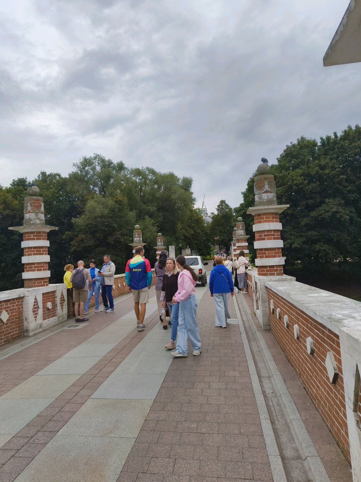 A group of people are walking on a stone bridge with brick railings in Tsaritsyno Park, Moscow, Russia. The bridge has decorative brick pillars topped with stone spheres and has a wide walkway with a patterned stone surface. The people are dressed in casual clothing, and some are looking at their phones. The weather is overcast, and the background shows lush green trees.