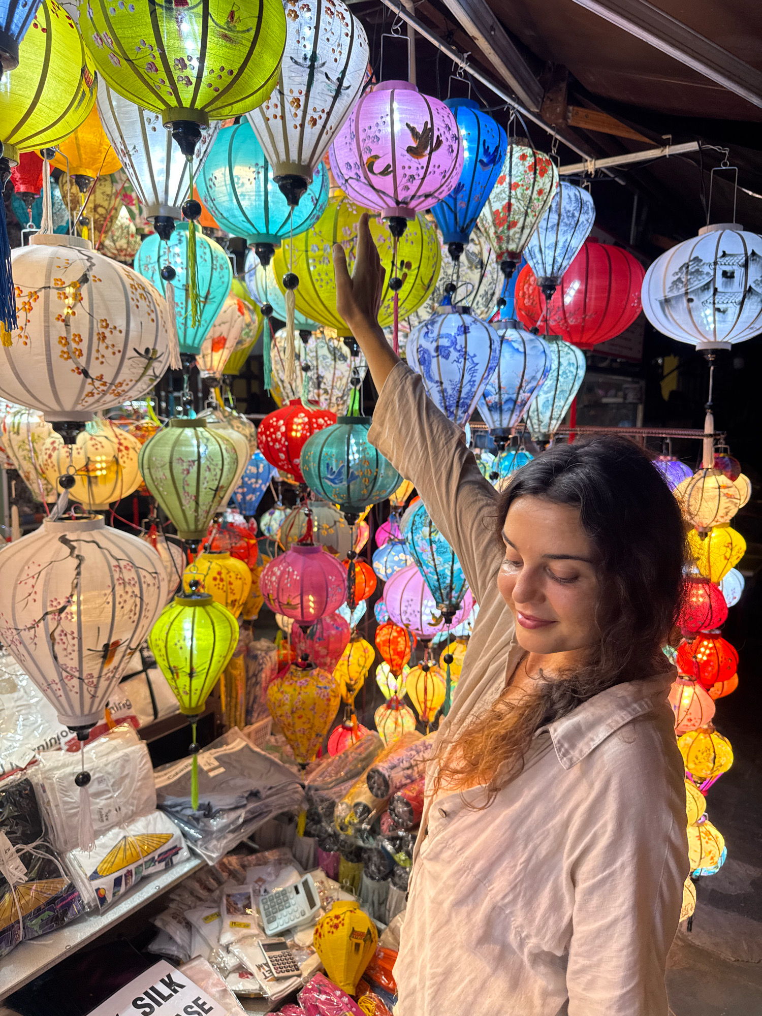 A young woman with dark, curly hair stands in front of a stall selling a large variety of colorful, ornate lanterns. She is wearing a light-colored collared shirt and her right arm is raised, her hand hovering near the lanterns. The lanterns are of various shapes and sizes, predominantly round or conical, and are decorated with intricate patterns, floral motifs, birds, and scenes of traditional architecture. The stall is brightly lit, with the lanterns themselves being the primary source of illumination, creating a vibrant and festive atmosphere. The background suggests an outdoor market setting, common in the historic town of Hoi An, Vietnam, known for its lantern-lit streets and traditional crafts. The woman appears to be looking down at the lanterns with a gentle smile, possibly considering a purchase. The scene captures a moment of cultural immersion and appreciation for the local artistry.