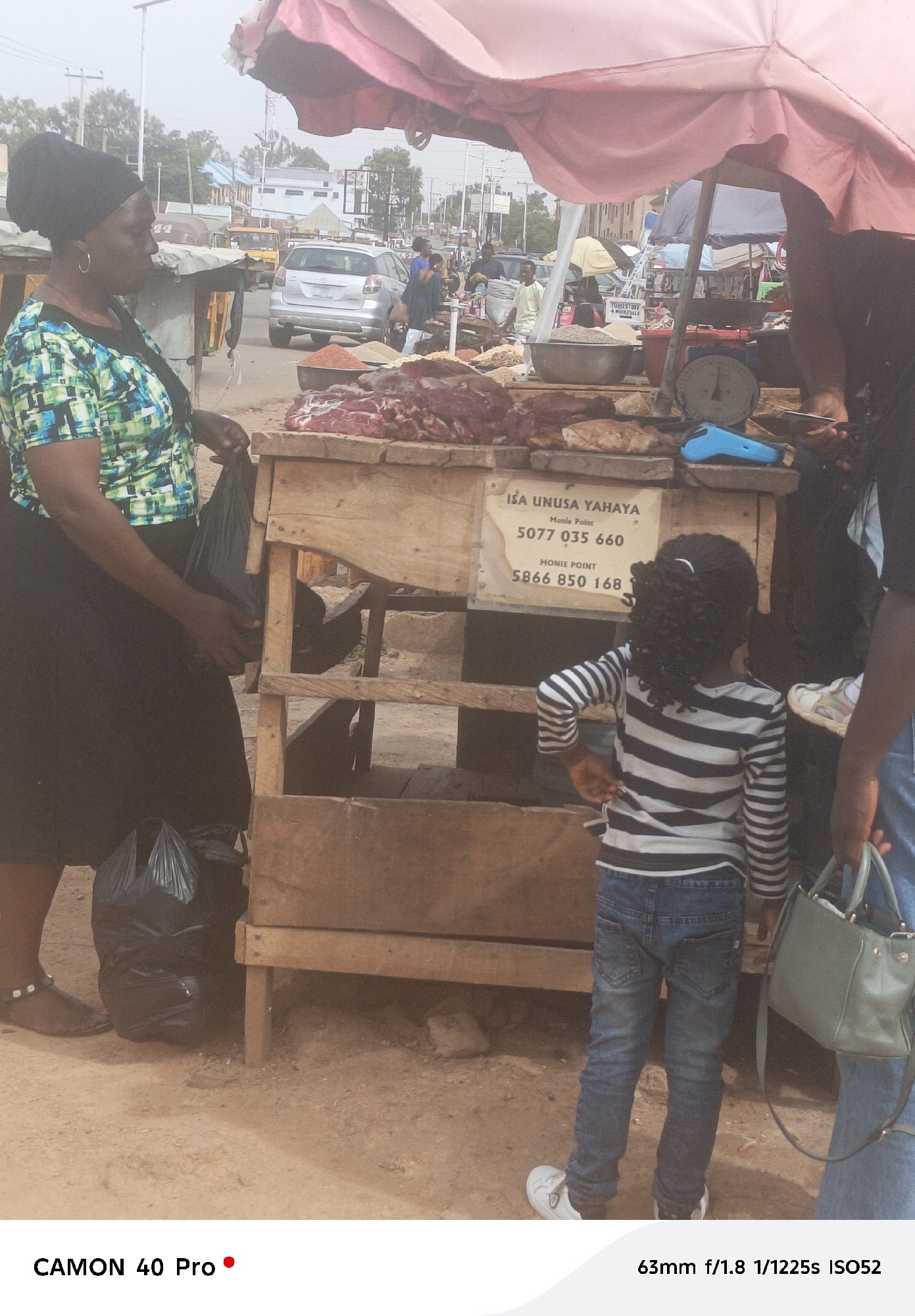 This image depicts a bustling market scene in Jos, Nigeria, centering on a meat vendor's stall. A large red umbrella provides shade over the stall, which is constructed from simple wooden planks. Prominently displayed on the counter are various cuts of raw red meat, alongside bowls of what appear to be spices or grains. A traditional weighing scale and a small blue device, possibly a mobile card reader, are also visible on the counter.
On the left, a woman dressed in a green and blue patterned shirt, a black skirt, and sandals holds a black plastic bag, seemingly interacting with the stall or waiting. In the right foreground, a young child with curly hair, wearing a striped long-sleeved shirt and blue jeans, stands facing away from the camera, observing the stall. Part of another adult, holding a light green handbag, is visible on the far right.
A sign on the wooden stall displays the name "ISA UNUSA YAHAYA" and two "Monie Point" mobile money contact numbers: "5077 035 660" and "5866 850 168". Another partially visible sign reads "FOODSTUF 4 WEBLESALE," indicating "FOODSTUFF FOR WHOLESALE." The background shows a busy street with vehicles and other market structures under a bright, hazy sky, typical of daytime in a warm climate. The ground is dusty.
The image was captured using a CAMON 40 Pro camera with specifications 63mm f/1.8, 1/1225s, and ISO52.