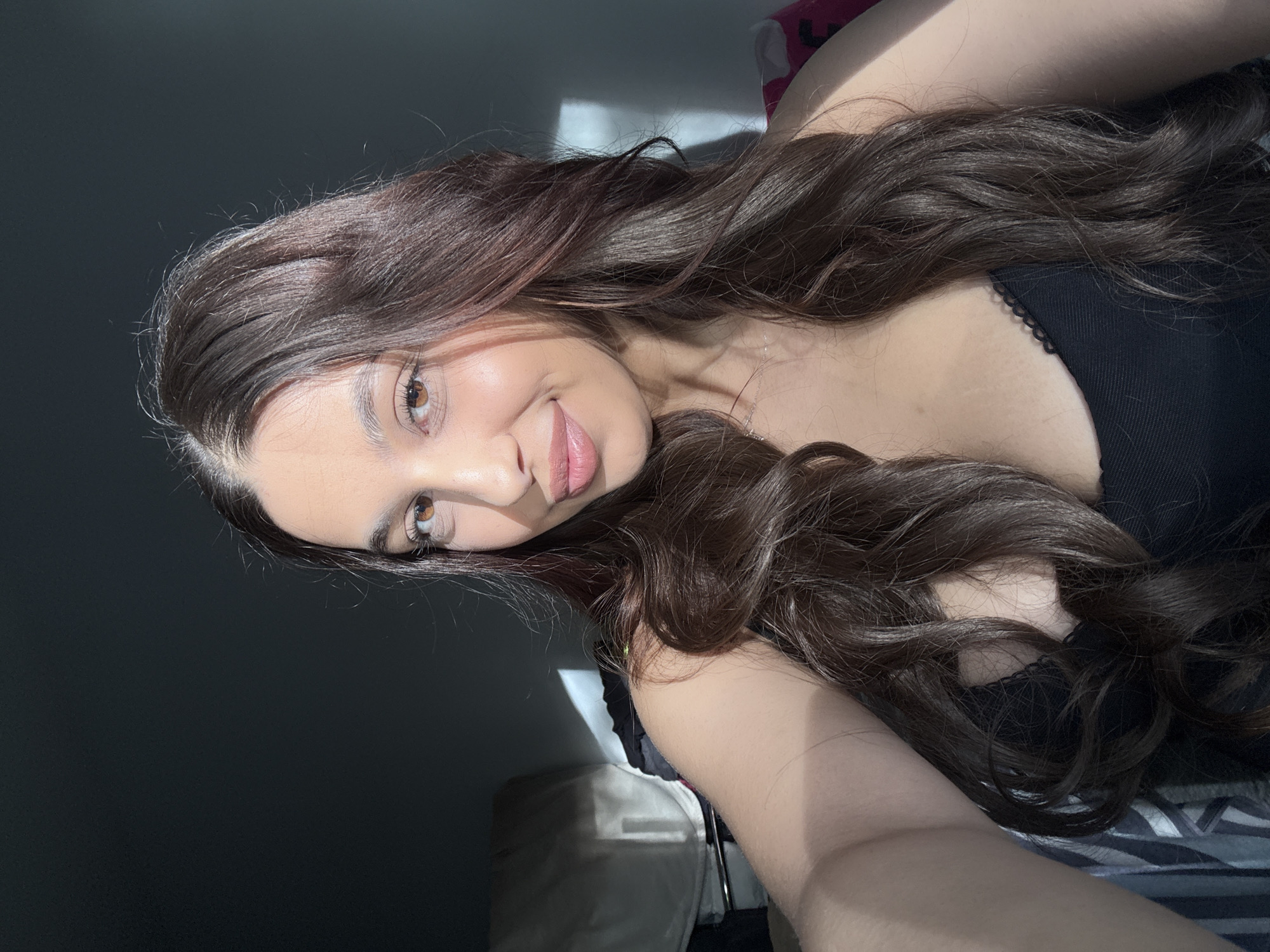 A young woman with long, wavy brown hair is lying down, looking at the camera with a slight smile. She is wearing a black top with lace trim. The lighting is bright and sunny on her face and hair. The background is partially a dark wall and partially bedding. There is no indication of the location being Mönchengladbach, Germany.