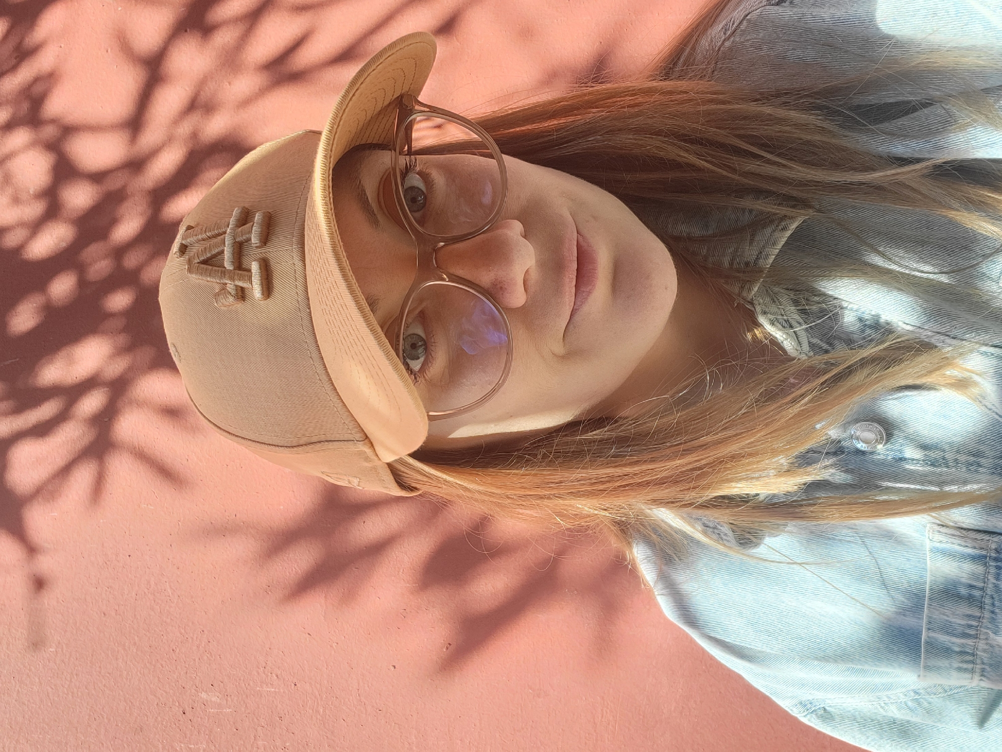 A person wearing a baseball cap and glasses is lying on a pink surface. The person has long, light brown hair and is wearing a light blue denim jacket. The baseball cap is light brown and has a raised logo on the side. Shadows from foliage are cast on the pink surface and the person's face. The location cannot be determined from the given image, therefore, the city of Budva, Montenegro cannot be confirmed.