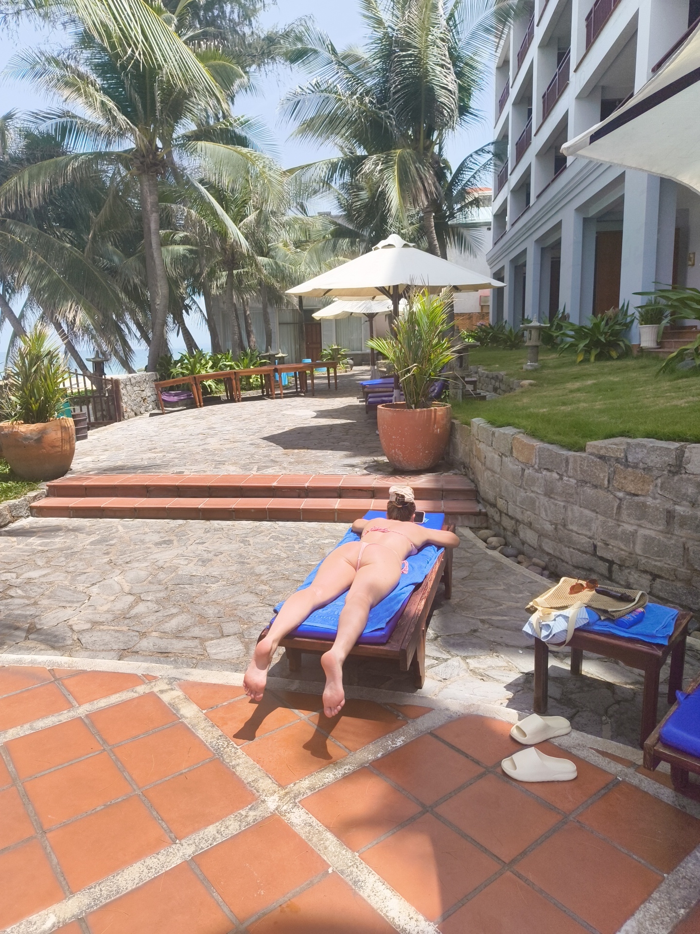 In the city of Phu Thuy Ward, Vietnam, a person is sunbathing on a lounge chair with a blue towel placed on top. The individual is wearing a light-colored bikini and is lying face down. The scene takes place outdoors, with a stone patio in the foreground and a pool area visible in the background. Lush palm trees and a hotel building are also present. There are several lounge chairs, a small wooden table with a straw hat, sunglasses, and a bottle of sunscreen, and a large potted plant. The ground is paved with a combination of stone and terracotta tiles.