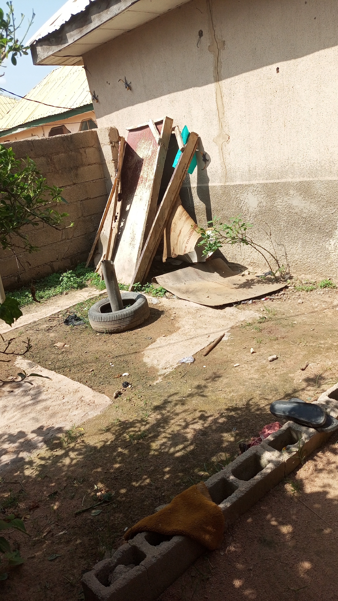 The image depicts an outdoor scene, likely a backyard or storage area, under bright daylight conditions. The setting is in Jalingo, Nigeria, as indicated by the provided context. In the background, a portion of a building with a pale, textured wall and a corrugated metal roof is visible. A concrete block wall forms a boundary to the left, with a glimpse of another building with a similar roof beyond it.
The central focus of the image is a collection of discarded materials leaning against the building. These include several large pieces of wood and what appear to be metal sheets, some with reddish-brown discoloration, suggesting they are old or rusted. A vertical pipe or pole is inserted into a worn tire that is lying on the ground. Several green objects, possibly pieces of fabric or plastic sheeting, are attached to the wooden supports.
The ground is a mixture of dirt and patches of dry grass. There is scattered debris on the ground, including a black plastic bag near the tire, and some white and brown pieces of litter further out. In the foreground, a concrete block structure runs horizontally across the frame. A piece of yellow-brown fabric is draped over one part of the blocks, and a black object, possibly a sandal, rests on another section. Shadows of foliage are cast across the ground, indicating the presence of trees or plants, with some greenery visible to the left of the frame and a small shrub growing near the discarded materials.
No people are visible in the image, and no specific activity or event is taking place. The scene appears to be one of general disarray and storage of old materials. The lighting suggests it is daytime, possibly mid-morning or afternoon, with clear skies. There is no visible text in the image.