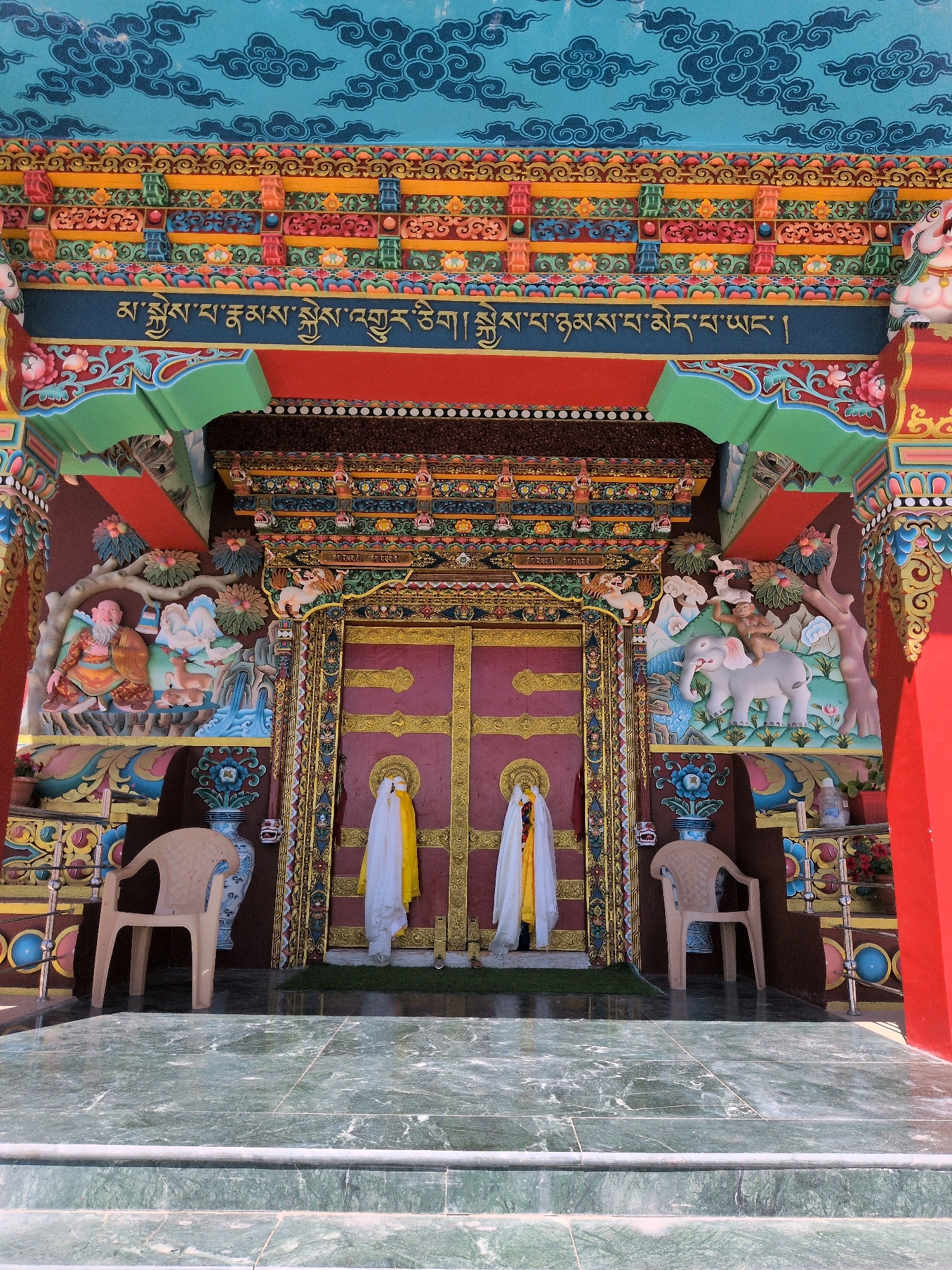 This image shows the ornate entrance to a Tibetan Buddhist monastery in Leh, India. The entrance is brightly painted in a variety of colors, including red, blue, yellow, and green, with intricate patterns and carvings. The central feature is a large, maroon door with gold detailing, flanked by white and yellow cloths draped over poles. On either side of the door, there are detailed relief carvings depicting scenes with figures, animals, and landscapes, characteristic of Tibetan Buddhist art. Above the door, a band of gold Tibetan script runs across a dark blue background. The entire structure is supported by elaborately decorated pillars and beams. In front of the entrance, two plain plastic chairs are placed on a green mat, and the foreground shows polished green marble steps. The overall impression is one of vibrant religious architecture, typical of monasteries in the Ladakh region.