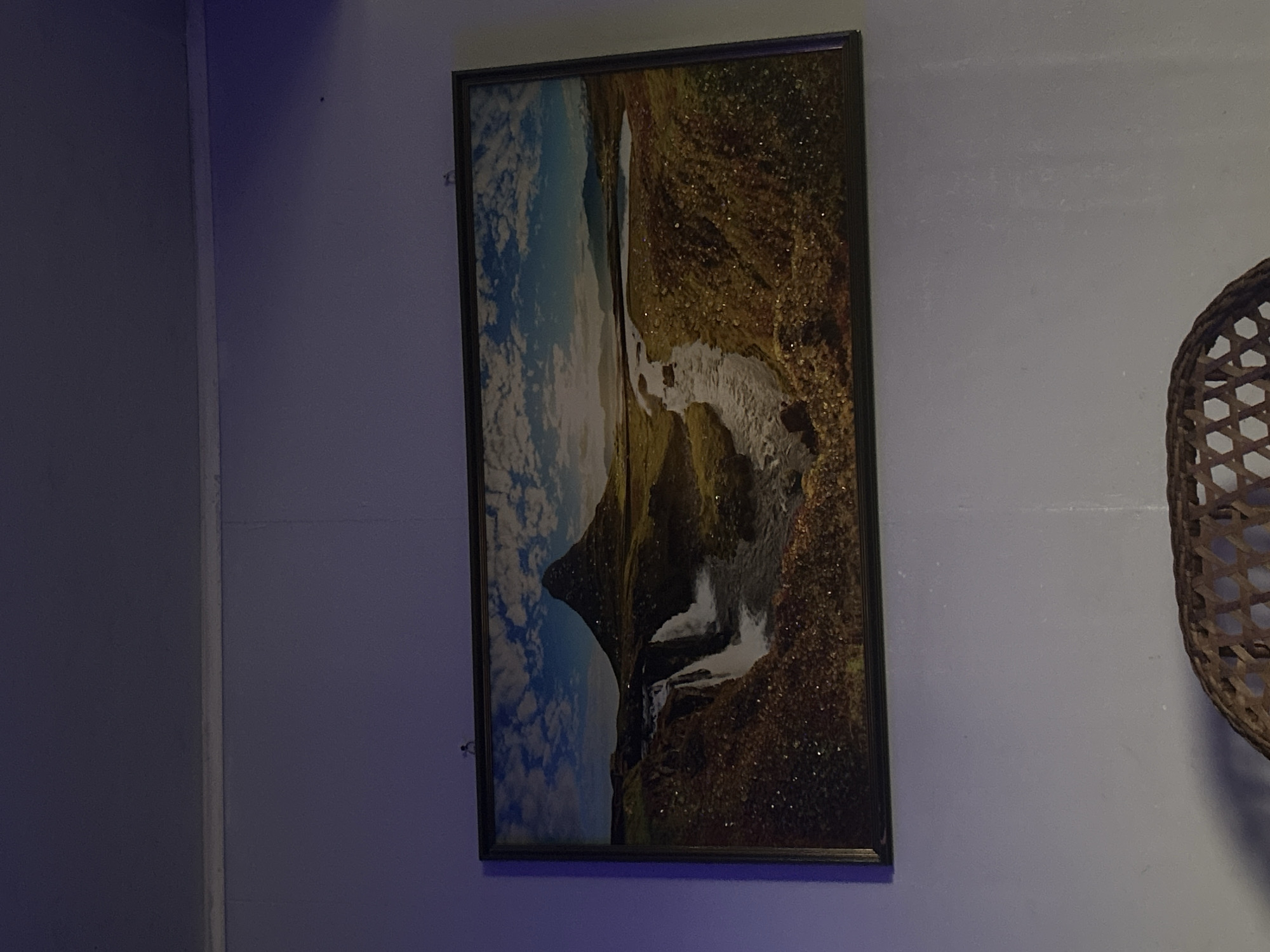 A framed landscape photograph is hung on a pale purple wall. The photograph depicts a scenic view of a mountain, a waterfall, and a river. The mountain is conical in shape and stands prominently in the background, bathed in sunlight. A waterfall cascades down a rocky cliff face on the left side of the image, feeding into a winding river that flows through a grassy, textured landscape. The sky is blue with fluffy white clouds. The photograph is bordered by a dark frame. The wall is illuminated by a soft, ambient light, possibly from a blue-toned lamp, casting subtle shadows. The bottom portion of the image shows a glimpse of a woven rattan object, likely a chair or basket, casting a distinct shadow on the wall. There is no indication of the city of Appany, Russia, in the image.