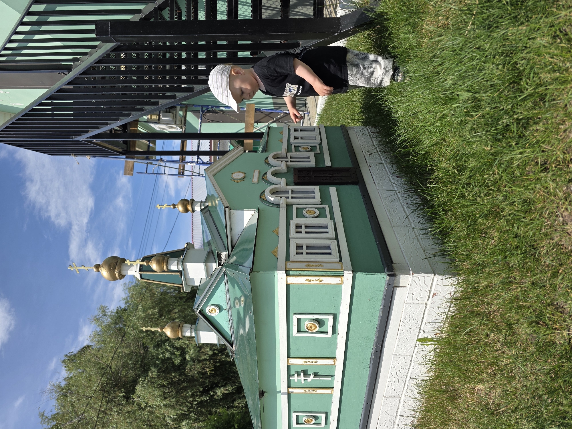 In Chaikovsky, Russia, a young child wearing a white cap, a black t-shirt, and camouflage pants stands on a patch of green grass, looking at a miniature replica of a building with green and white details and golden domes. The replica appears to be a church or chapel. To the right of the miniature building, a black metal staircase ascends along the side of a larger, pale green building. The sky above is blue with scattered white clouds. The scene is outdoors, with trees visible in the background.