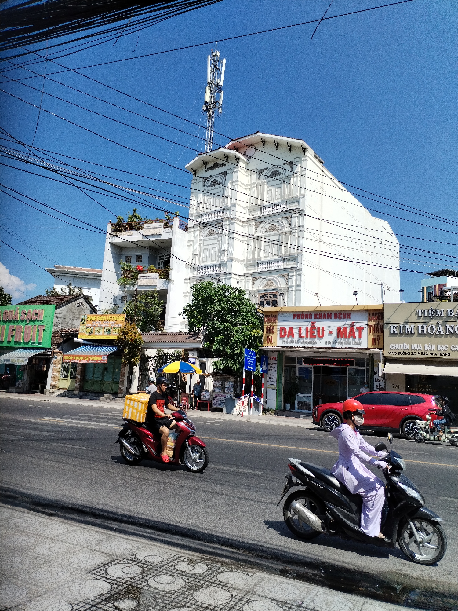 This media file depicts a street scene in Bac Nha Trang Ward, Vietnam, under a clear blue sky, suggesting a sunny day. The focus is on the street traffic and buildings. Two individuals on scooters are prominent. The first, a person in a black t-shirt and shorts, is riding a red scooter with a large yellow cargo box attached to the back. The second person, wearing a light purple outfit and a red helmet, rides a black scooter.
The background features a multi-story white building with ornate architectural details, resembling classical design elements. Other buildings line the street, some with awnings and signage, indicating commercial establishments. Visible text includes "SHOP FOOD LE THANG" and names of businesses like "DA LIEU - MAT" and "TIEM BAC KIM HOANG". A red car is parked on the side of the road. The presence of numerous power lines overhead is a notable detail of the urban landscape. The scene captures a typical day with everyday activities like transportation and commerce.