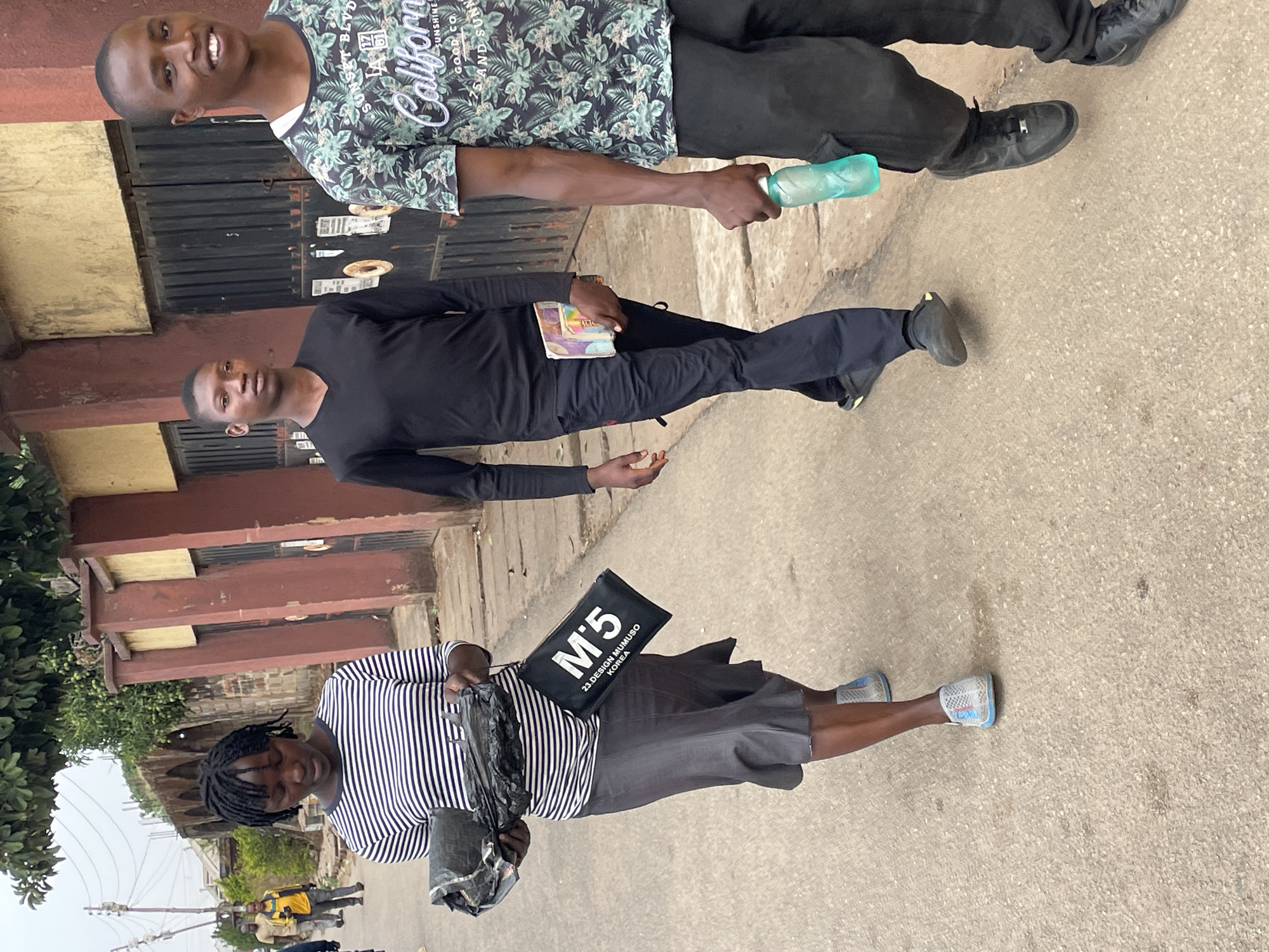 Three individuals are standing on a paved surface, likely a street, in Jos, Nigeria. The person closest to the viewer is wearing a striped shirt, a grey skirt, and light-colored sneakers. They are carrying a black tote bag with "M'5" printed on it and a black plastic bag. The middle person is dressed in all black and is holding a book or pamphlet. The third person wears a shirt with a leafy pattern and dark pants and holds a water bottle. The background includes a building with brown and yellow walls and barred windows. Green foliage is visible to the side, and there are other people further down the street. The lighting suggests it is daytime.