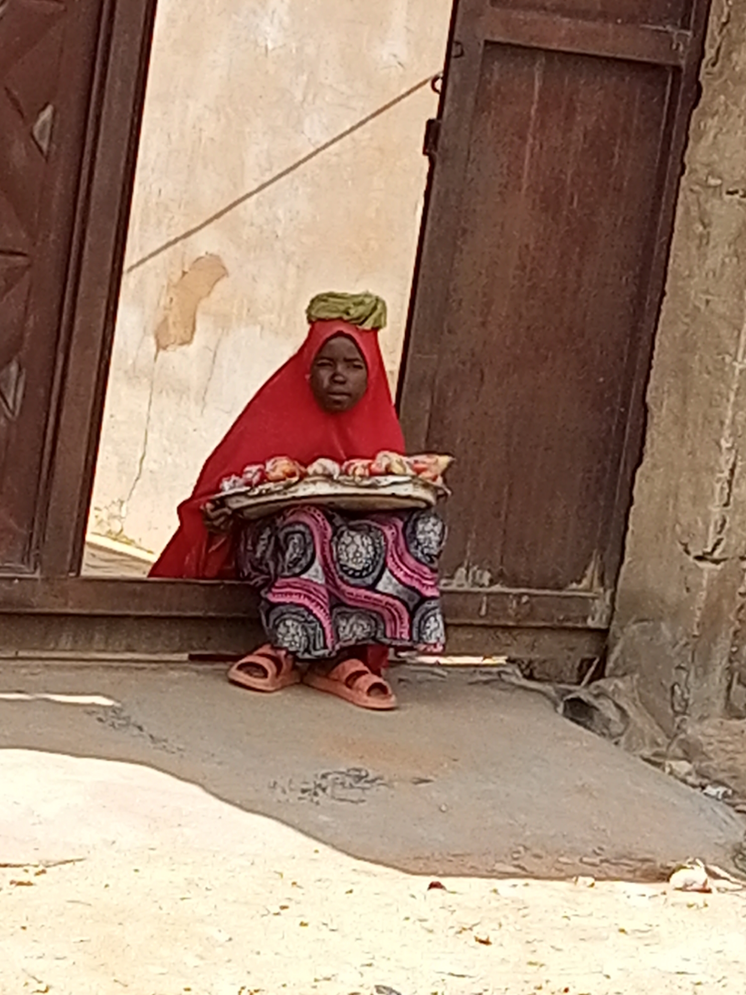 A young person, dressed in a red hijab and a patterned skirt, sits on a step in front of a weathered wooden door. They wear simple sandals and hold a tray of food, possibly fruits, on their lap. The background shows a textured, light-colored wall and a dark, aged wooden door. The ground in front of them is sandy and uneven, with some debris scattered around. The overall scene appears to be outdoors in a somewhat rural or older urban setting in Gombe, Nigeria.