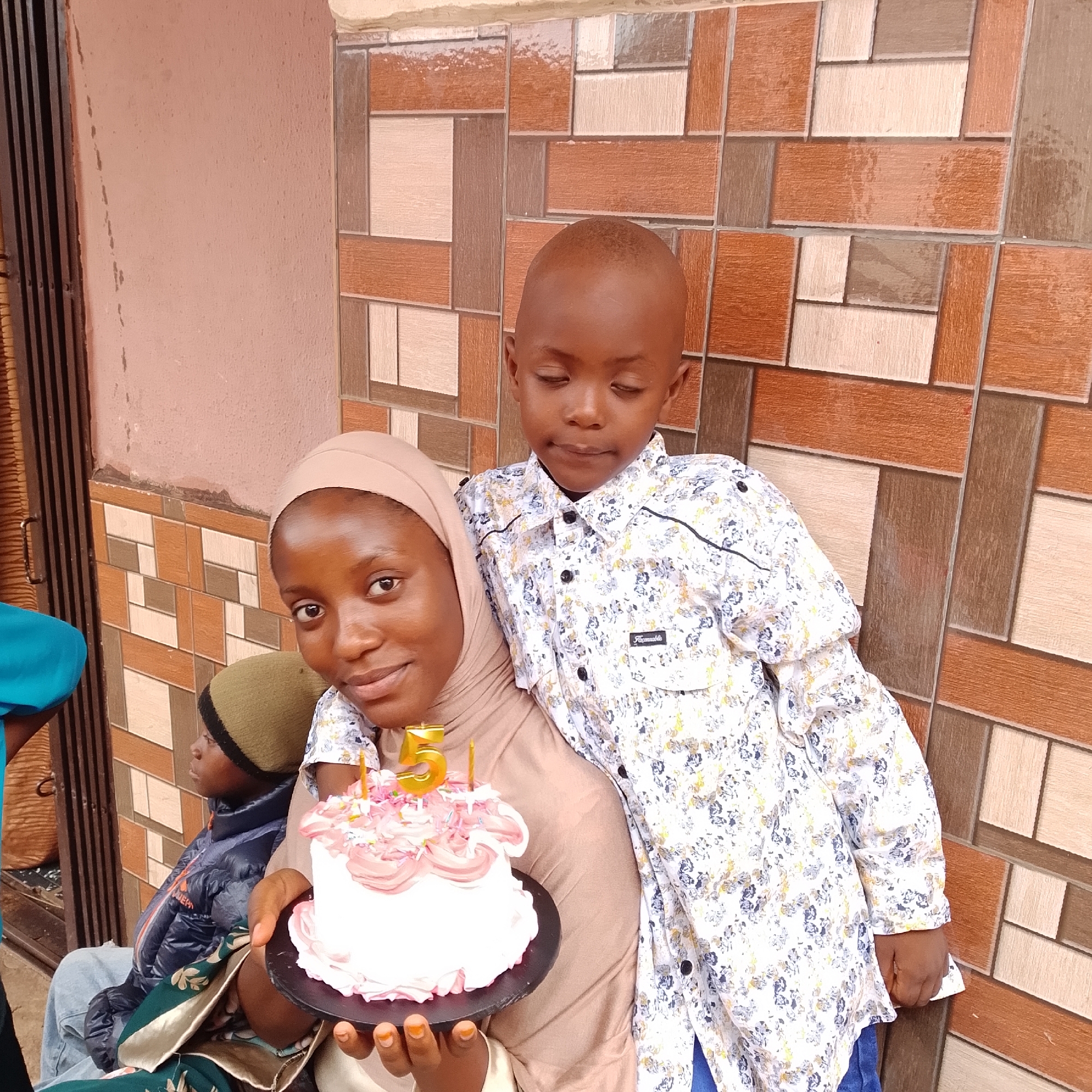 A medium shot photograph shows a woman wearing a light brown hijab, a young boy in a floral shirt, and another child in a jacket and beanie. The woman is holding a white birthday cake with pink frosting and a gold number "5" candle. She is smiling and looking towards the camera. The young boy is standing behind her, leaning forward with his eyes closed. The other child is sitting in the background, looking to the left. The scene appears to be outdoors, in front of a wall with a patterned tile facade. The background wall has a textured, reddish-brown and beige tile pattern. To the left, a portion of a dark metal gate or door is visible. The lighting suggests it is daytime.