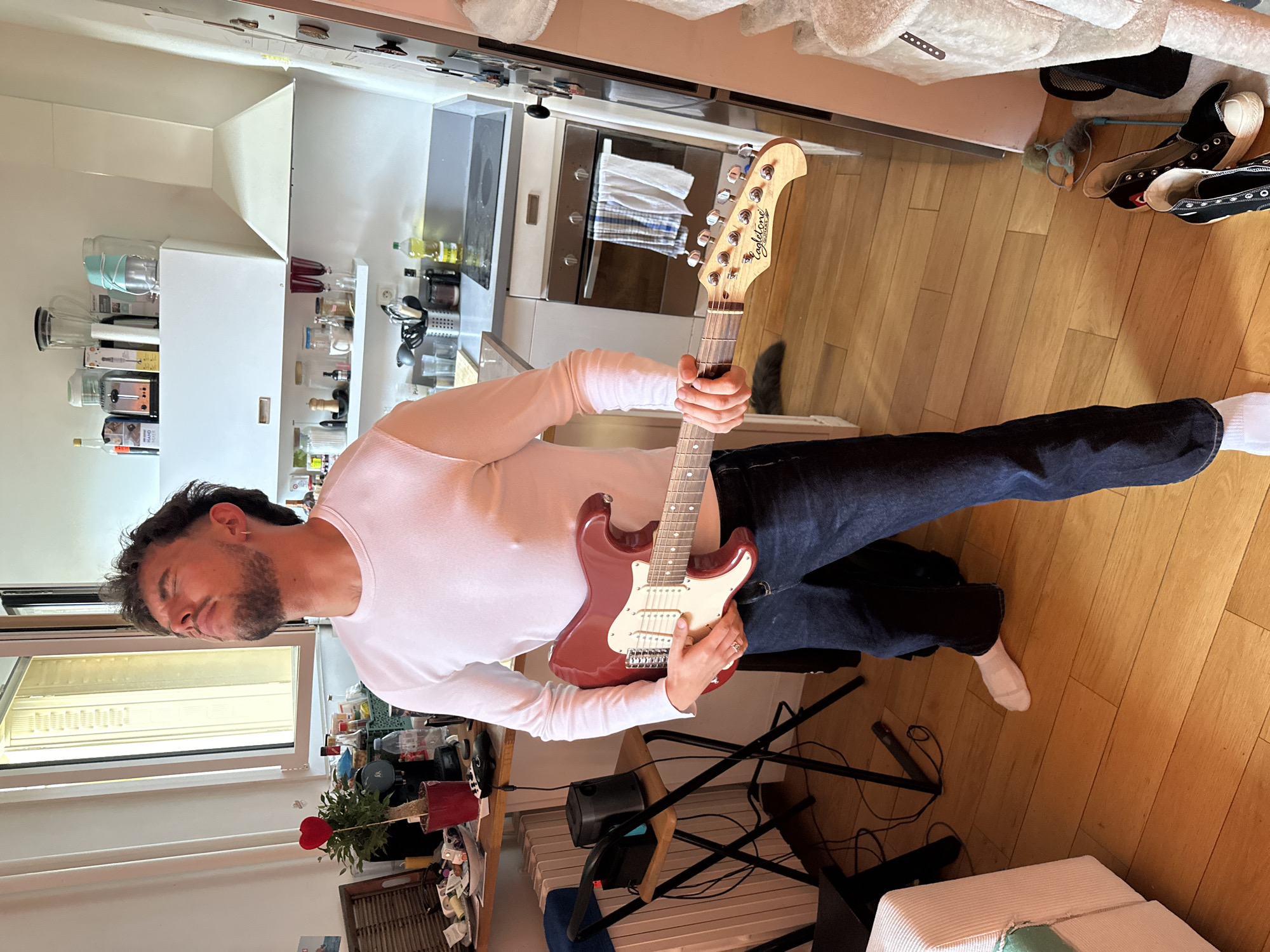 A man is captured playing an electric guitar inside what appears to be a home in Paris, France. He is positioned in the foreground, holding a dark red electric guitar with a white pickguard. The man wears a white long-sleeved shirt, dark blue jeans, and white socks, with a ring visible on his right hand. He has dark, curly hair and a short beard, and his gaze is directed downwards towards the guitar as if in concentration.
The setting is an indoor space that blends a kitchen area with a living or casual space. In the background, to the left, is a clean, modern kitchen featuring white cabinets, stainless steel appliances like a stove and oven, and a range hood. The kitchen counter is adorned with various items including bottles of oil, jars, and kitchen gadgets, one box partially showing the text "HAND MIXER." Above the stove, a white and blue checkered towel is draped over an oven handle.
To the right, beyond the man's feet, a wooden floor extends, with a pair of black high-top sneakers (possibly Converse) and a small, light-colored object resembling a mop or duster lying nearby. A fluffy white blanket or rug is also visible. In the bottom left, a window provides natural light, and a small potted plant with a red heart decoration sits on a surface. The guitar's headstock clearly displays the brand name "Eagletone." The overall impression is a casual, daytime scene in a lived-in environment.