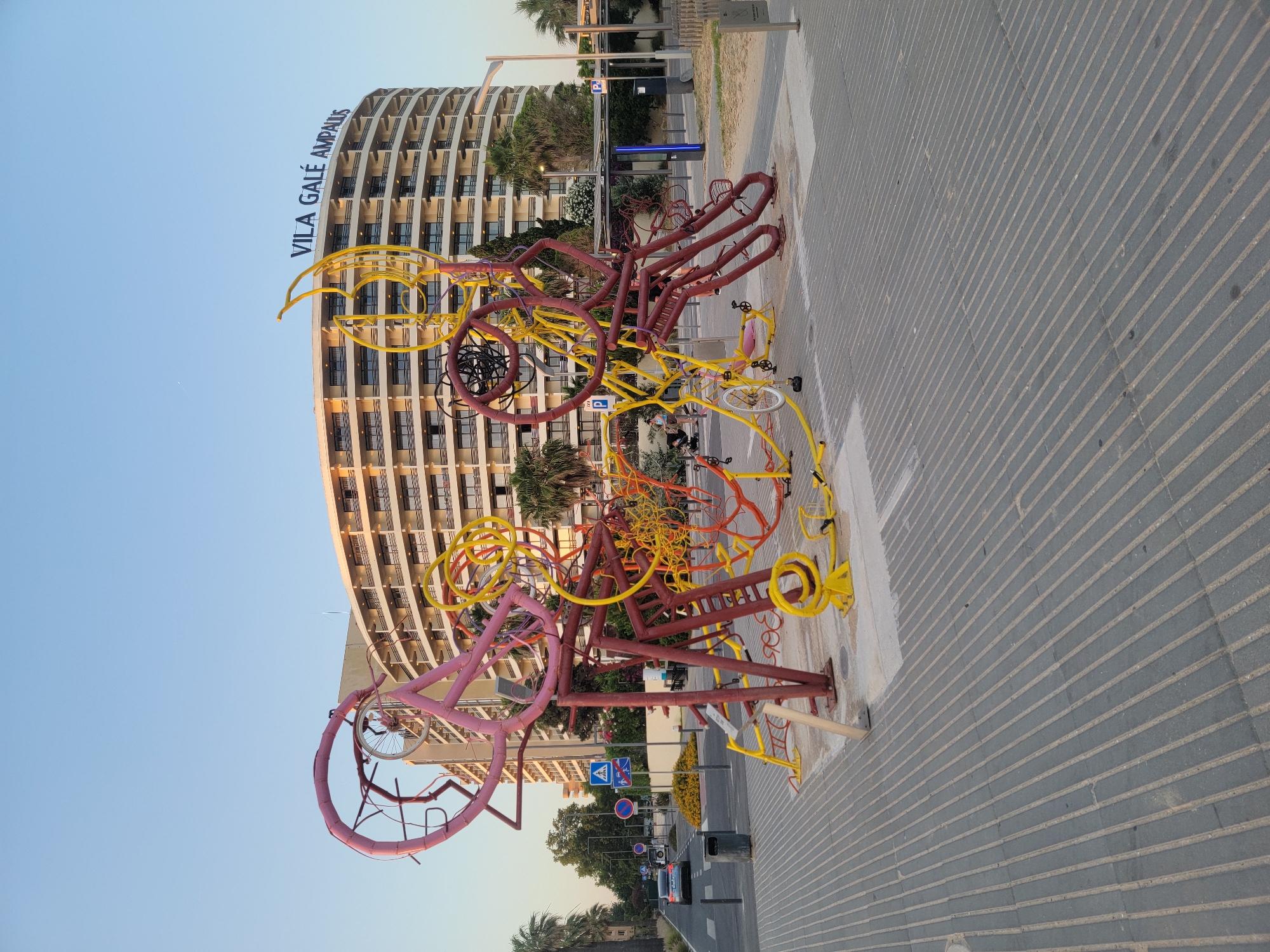 The image shows a large, abstract sculpture made of colorful metal tubes, primarily red, yellow, and pink, resembling intertwined bicycle parts and abstract forms. The sculpture is situated on a paved walkway with a tactile paving pattern. In the background, a large, curved, multi-story building with a light-colored facade and dark balconies dominates the scene. The building has a sign at the top that reads "VILA GALÉ AMPALUS". Palm trees and other greenery are visible around the building and along the street. A street with cars, traffic signs, and a trash can is present to the left of the sculpture. The sky is clear and a light blue, indicating daytime, likely late afternoon or early evening due to the lighting. The location is Quarteira, Portugal.