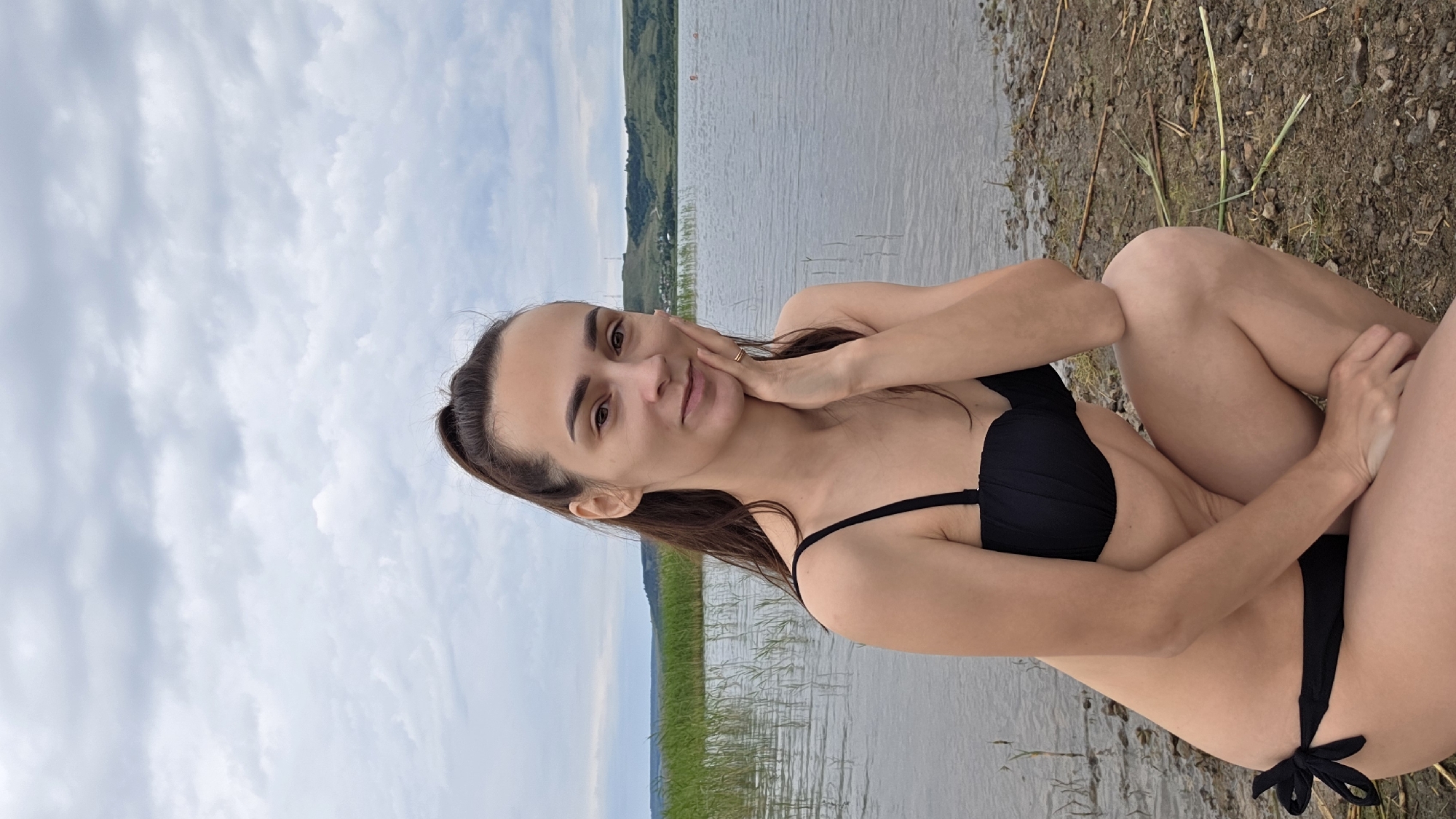 A woman wearing a black bikini crouches on a gravelly shore next to a body of water. She has long brown hair pulled into a ponytail and rests her left hand on her cheek. The background shows a calm lake with reeds along the edge, rolling green hills, and a cloudy sky. There are two indistinct figures in the water further from the shore.