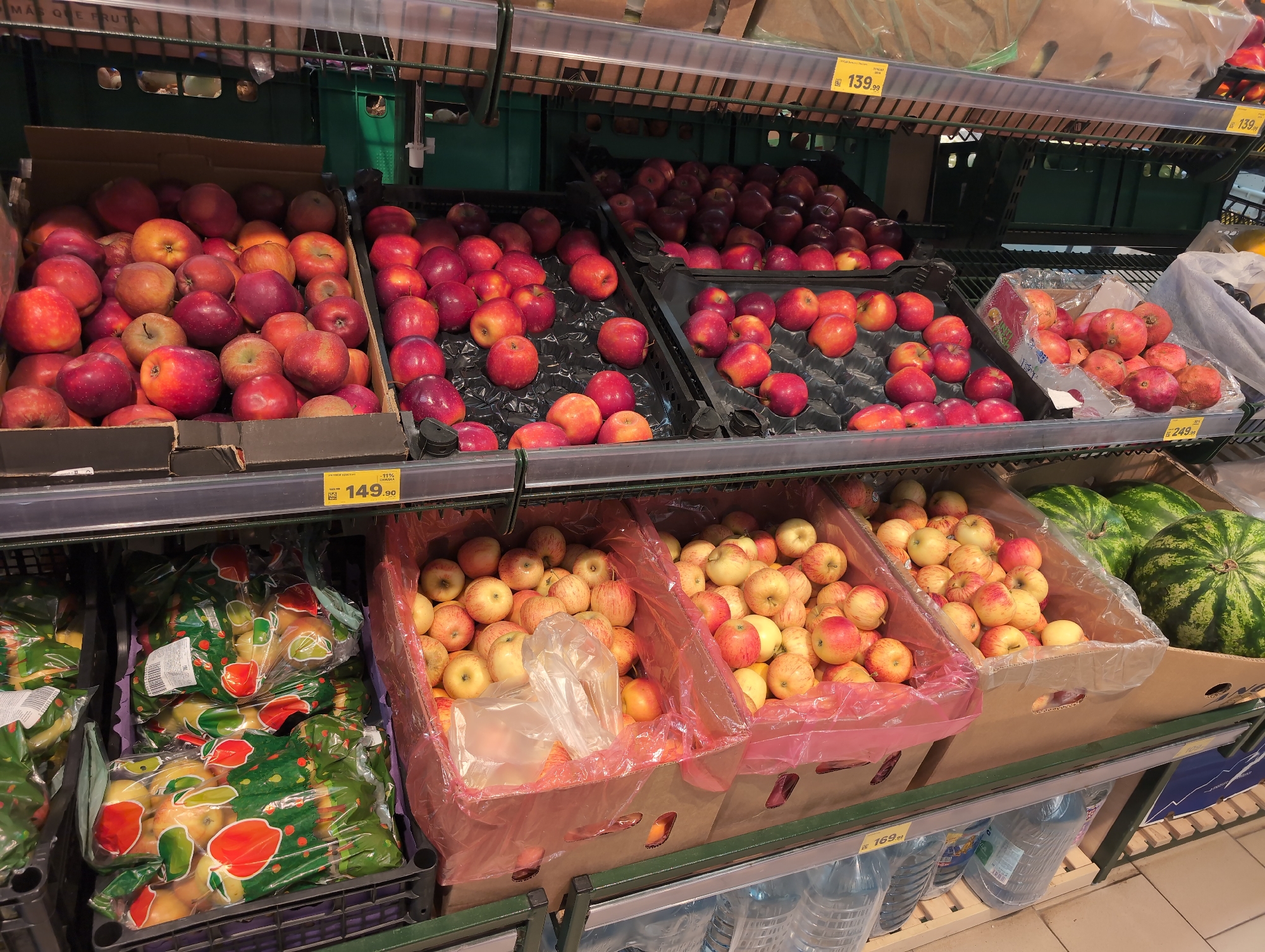 This image shows a display of fruit in a supermarket in Krasnodar, Russia. The fruit is arranged on shelves, with apples in cardboard boxes and plastic bags, and watermelons in a cardboard box. The apples are red and yellow, and the watermelons are green. There are also price tags visible on the shelves, indicating the cost of the fruit. The supermarket appears to be well-stocked, with a wide variety of produce on display. The lighting is bright and even, suggesting that the image was taken during the day. The overall impression is one of a clean and organized supermarket, with a focus on fresh produce.