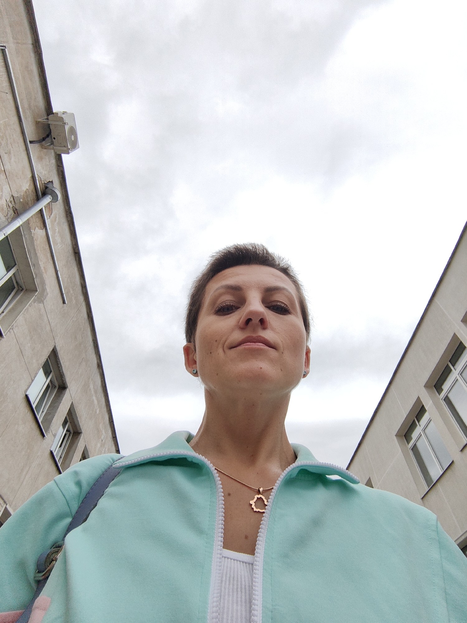 A low-angle, full-body shot shows a woman standing between two buildings under a cloudy sky, possibly in Minsk, Belarus. The woman, with short brown hair and light skin, wears a mint green zippered jacket over a white tank top. A rose gold necklace with a hedgehog pendant is visible around her neck. To her left, a cream-colored building features a window and an outdoor air conditioning unit mounted on its side. To her right, a lighter cream-colored building displays several windows. The overall impression is of an urban setting.