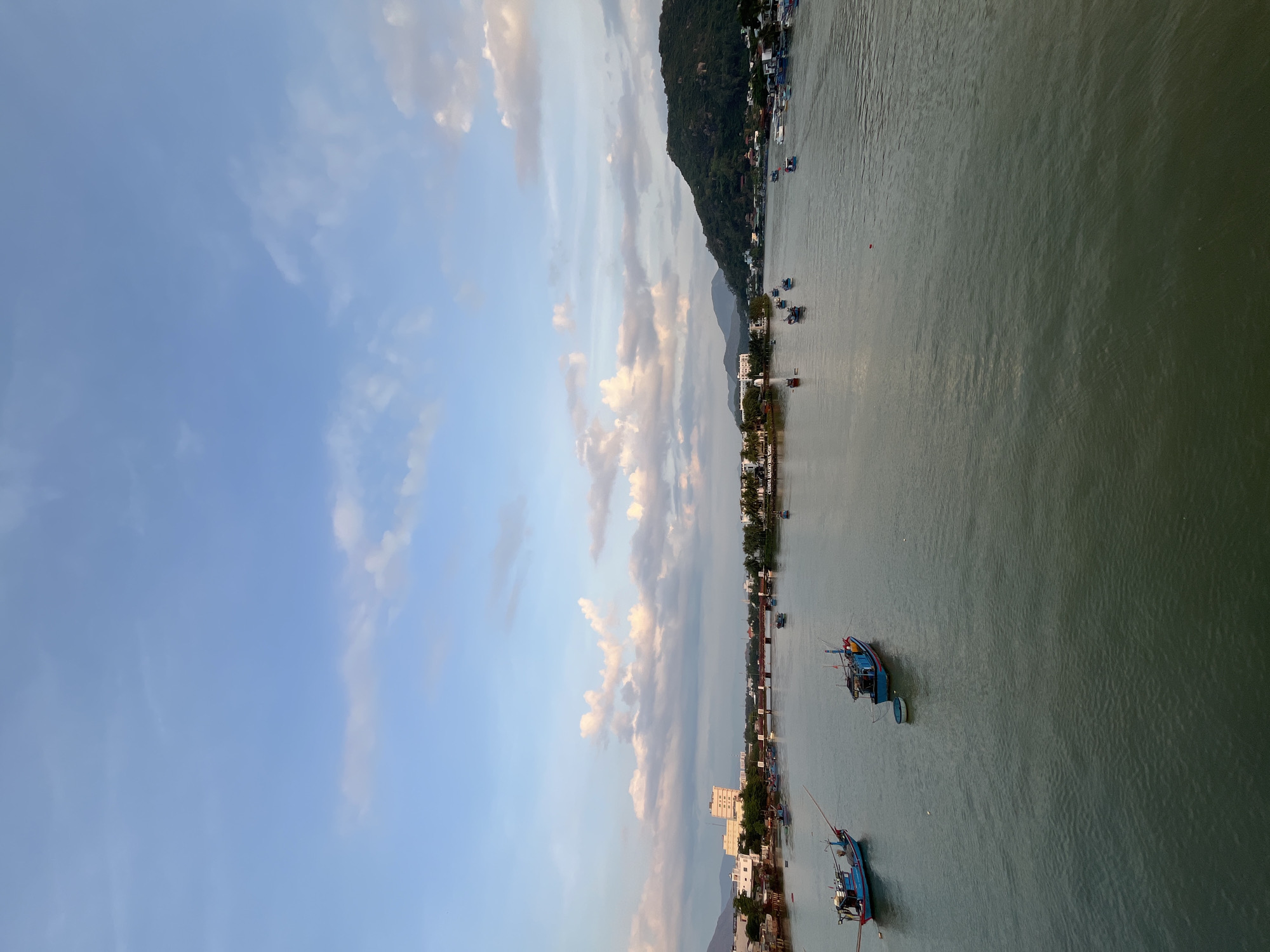 The image is a long shot of a body of water with buildings and land on either side. There are boats in the water, and a mountain is on one side of the body of water. The sky is blue with clouds. This image contains the city of Bac Nha Trang Ward, Vietnam.