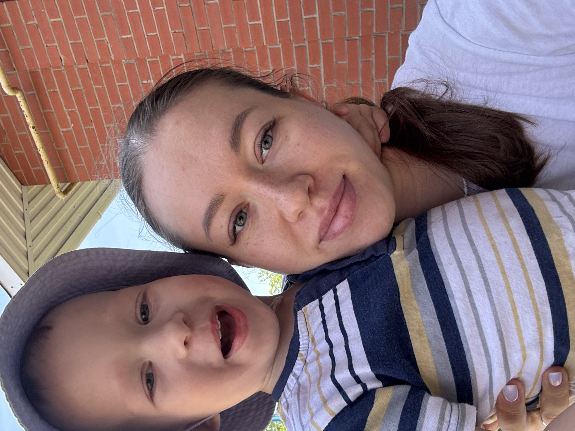 This is a selfie featuring a woman and a child, possibly taken in Ферма, Russia, based on the request. The woman has light skin, brown hair pulled back, and is smiling slightly at the camera. The child, who is wearing a light gray hat and a striped shirt with navy blue, yellow, and white stripes, is looking at the camera with their mouth open, seemingly mid-speech. The background reveals a brick wall and what appears to be the underside of a roof structure with yellow supports. The photo is taken at an angle, with the camera pointing upwards.