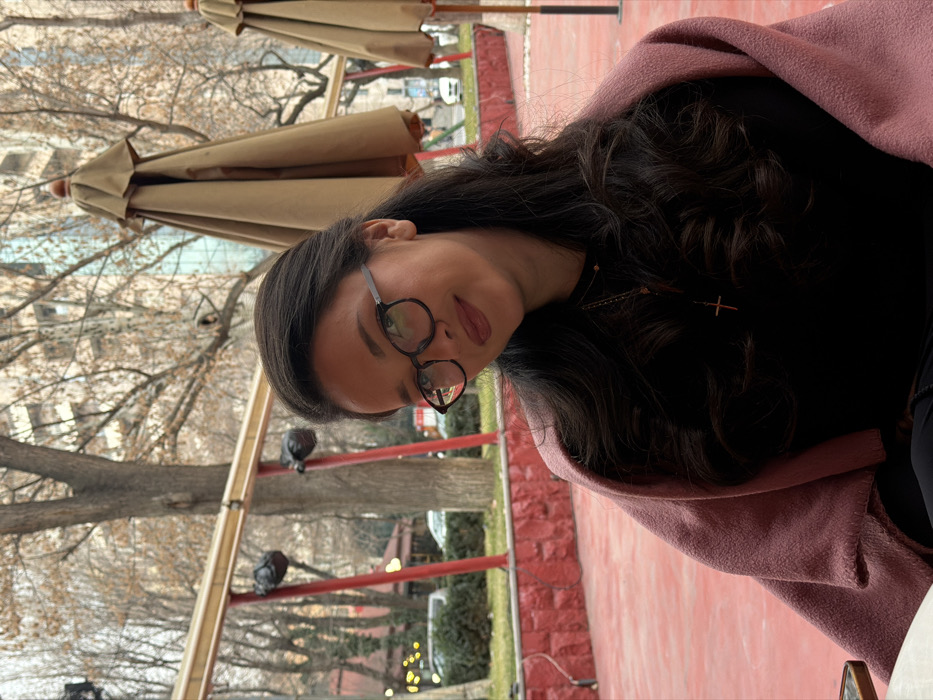 A woman with dark hair and round glasses poses outdoors. She wears a pink wrap or sweater over a black top. A necklace with a small cross is visible. The background shows a park or outdoor cafe setting with trees, closed umbrellas, and red brick elements. The scene appears to be in daylight with a somewhat overcast sky.