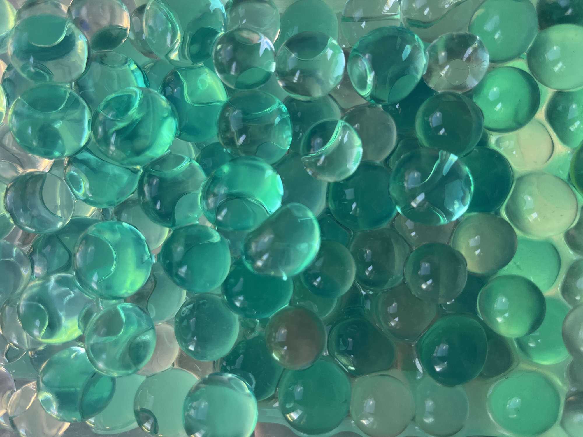 The image is a close-up, overhead view of a collection of clear, gelatinous spheres in varying shades of green. The spheres appear to be water beads, also known as Orbeez. They are densely packed together, creating a textured surface. The lighting creates highlights and reflections on the smooth surfaces of the spheres, giving them a glistening appearance. Some spheres are more translucent than others, allowing a glimpse of the internal structure or perhaps other spheres beneath them. The color palette ranges from a pale, almost clear green to a deep, emerald green. The arrangement of the spheres is somewhat organic, with some appearing perfectly spherical and others slightly distorted due to the pressure from their neighbors. There is no visible text in the image. The context suggests a still life or macro photography subject, focusing on the visual appeal of these translucent orbs and their play with light. No specific time of day, weather, or location cues are discernible. The overall impression is one of a vibrant, cool, and possibly tactile display.