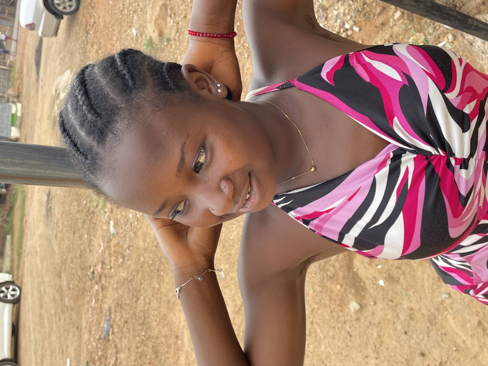 A portrait-style image, taken outdoors in Barakin Akawo, Nigeria, features a young Black woman looking gently to her left with a subtle, pleasant smile. She has dark skin and her hair is styled in neat, dark braids. Her eyes are a striking light brown or amber color. She wears a nose piercing, two small stud earrings in her visible ear, a delicate gold chain necklace with a small bead, and two bracelets – one made of red beads and another silver chain bracelet with small charms.
She is dressed in a vibrant sleeveless top or dress featuring an abstract pattern of bold pinks (dark and light), black, and white. Her hands are positioned near her head, with elbows possibly resting on a surface, a dark bar of which is visible in the foreground.
The background, largely out of focus, shows an unpaved, sandy-brown ground with scattered small debris. A white car is partially visible in the distance on the left side, suggesting an open area or residential street. The lighting indicates it is daytime, with soft, natural illumination on her face. No visible text is present in the image.