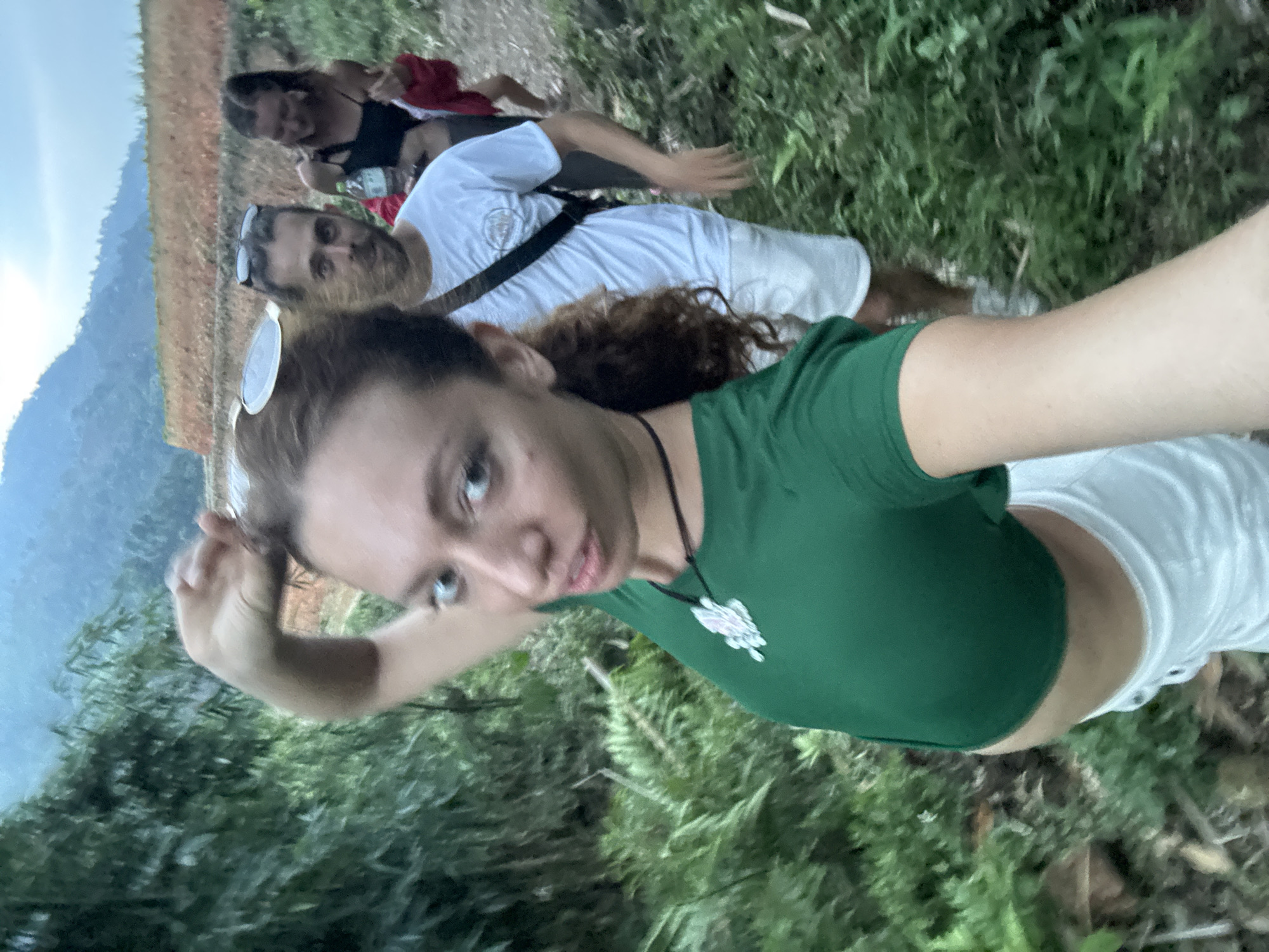 The image is a vertical-oriented selfie taken outdoors in Nậm Dịch Commune, Vietnam, featuring three individuals against a natural, mountainous backdrop.
In the foreground, a woman with curly brown hair and light eyes is taking the photograph, looking directly at the camera with a slight expression. She wears a dark green short-sleeved crop top with a decorative white pendant on a black cord necklace, and white athletic shorts. Her left arm is bent, holding the camera.
Behind her, a man with a beard and sunglasses resting on his head stands, also looking towards the camera. He is wearing a white t-shirt with a faint circular graphic on the chest and light-colored shorts. A black strap, likely from a bag, crosses his body. Further in the background, a third person is partially visible, sitting or kneeling amidst the vegetation, wearing a dark top and red shorts, with a green water bottle nearby.
The setting is characterized by abundant green foliage, including bushes and ferns, suggesting a lush, natural environment. Areas of reddish-brown soil or earth are visible. In the distant background, a large, hazy mountain range rises under a bright, light blue sky, indicating clear daytime weather. The scene conveys a sense of outdoor activity, possibly hiking or exploring, against a scenic Vietnamese landscape. No specific text is clearly legible on any clothing or objects.