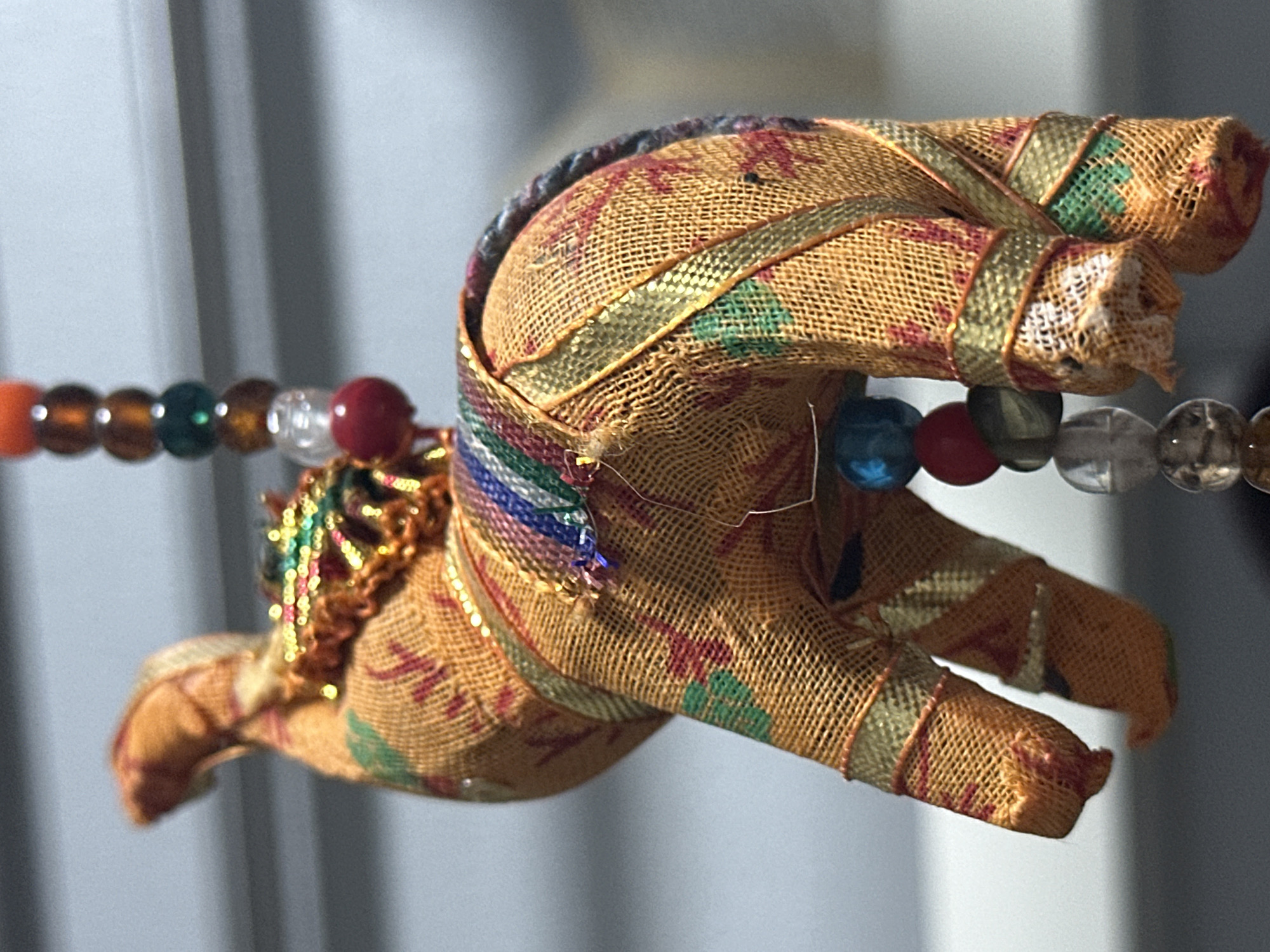 An orange fabric elephant ornament with colorful beaded trim hangs against a blurred background of gray horizontal lines. The elephant is adorned with gold and multi-colored striped ribbons. A strand of beads, including red, green, and brown spheres, hangs below the elephant. The ornament appears to be a souvenir or decorative item. The location is identified as Nevinnomyssk, Russia.