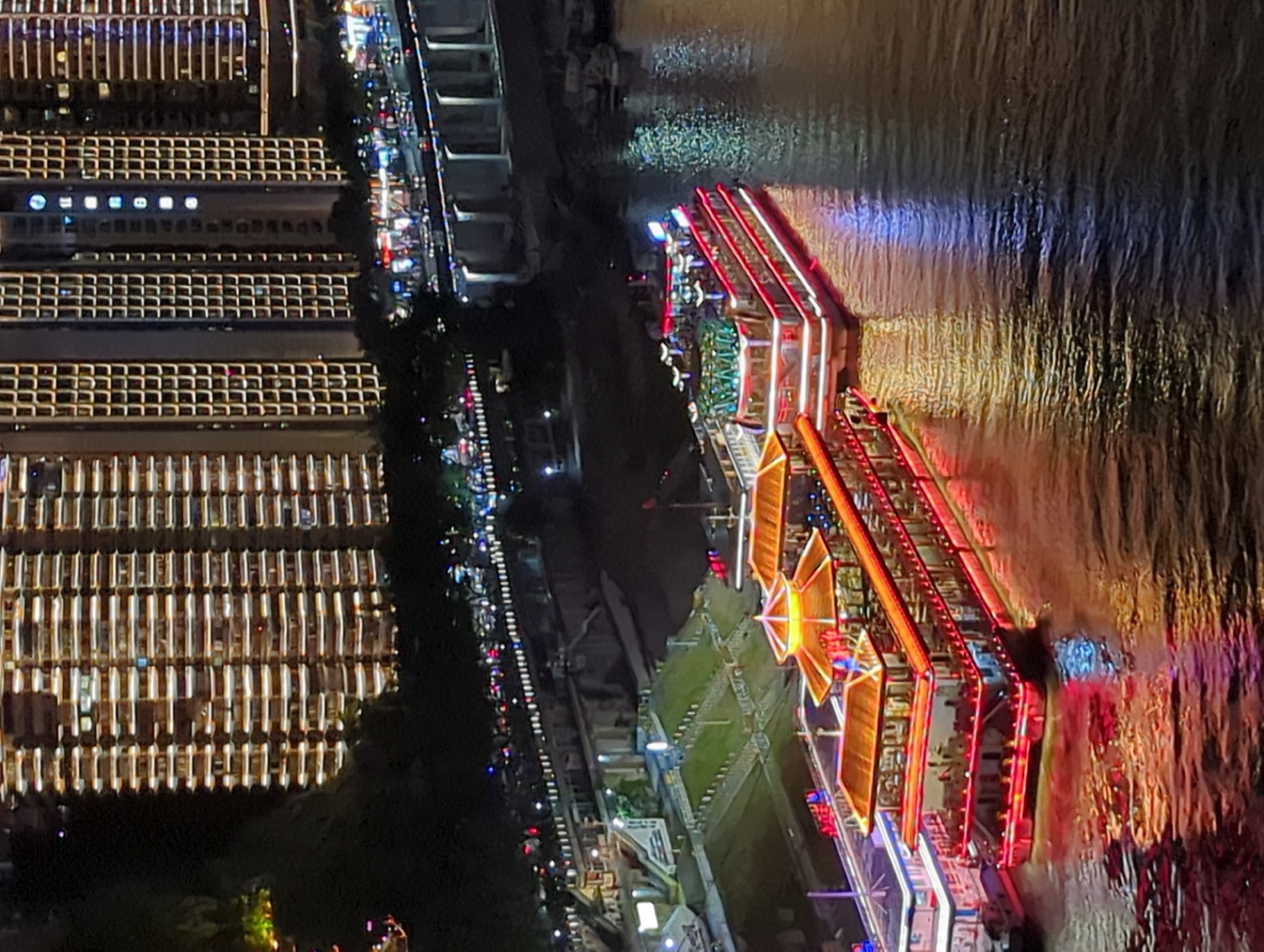 At night in Yuzhong District, China, two illuminated tour boats are docked side-by-side on a wide river. The boat in the foreground has multiple decks, with brightly lit windows and red and white lights outlining its structure. Its roofs are ornately decorated with glowing yellow and orange elements. The second boat, to its right, is similarly lit with red and blue lights, and its multi-level structure is visible. The water reflects the vibrant lights of the boats and the distant city. In the background, a densely packed cityscape of tall buildings with numerous illuminated windows rises up, indicating a bustling urban environment. A bridge with illuminated supports spans the river in the middle ground, and a tree-lined promenade with streetlights is visible along the riverbank. The overall scene captures the energetic nightlife of a waterfront city.