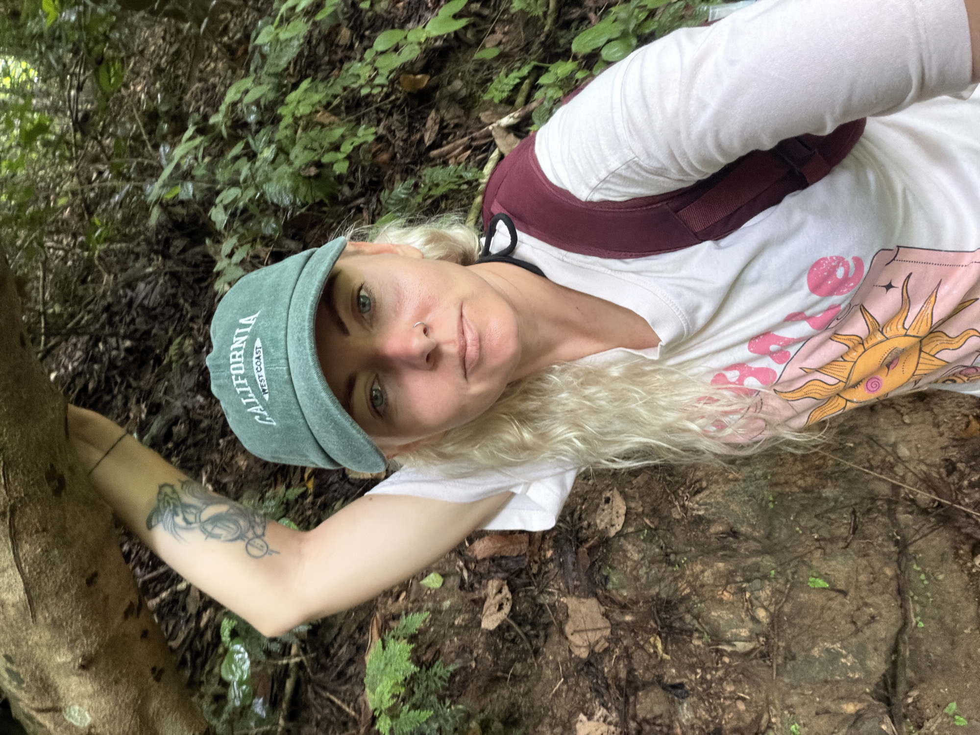 A young Caucasian woman with long blonde hair and a tattoo on her left arm is taking a selfie in a forest. She is wearing a green baseball cap with "CALIFORNIA WEST COAST" embroidered on it, a white long-sleeved t-shirt with a sun graphic, and a maroon backpack. She is holding onto a tree trunk for support, and the background is filled with lush green foliage and trees. The ground is covered in dirt, rocks, and fallen leaves. The image appears to be taken in a natural, outdoor setting, possibly a hiking trail. There is no specific indication of the location being in the city of Thapene, Laos.