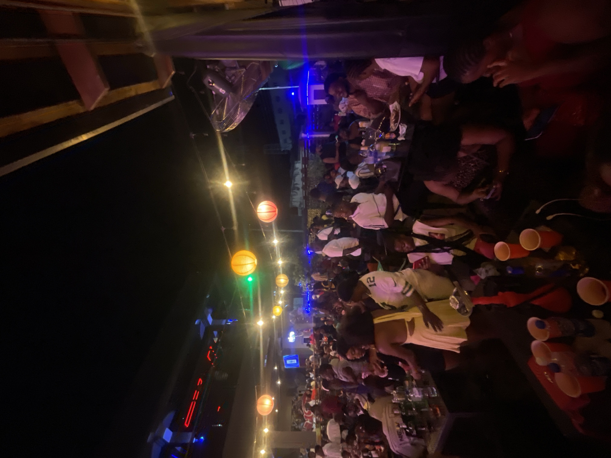 The image shows a crowded outdoor bar in Lokoja, Nigeria, at night. Patrons are seated at tables, with cups and bottles visible on the surfaces. The people are predominantly Black. Overhead, orange and yellow spherical lights illuminate the scene, with cooler blue lights in the distance. A large fan is visible overhead, along with some structure.