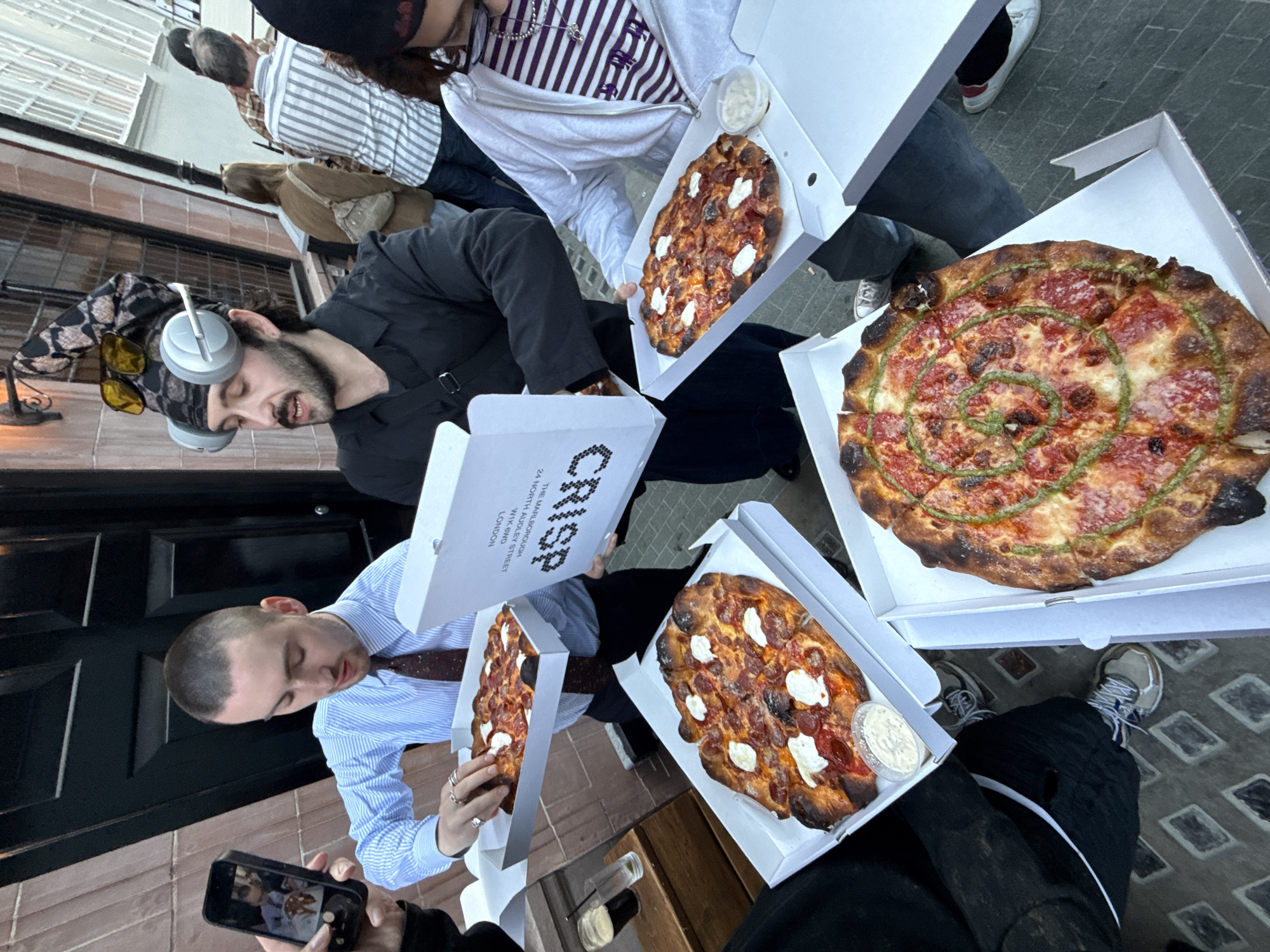 The image captures a casual street scene in London, United Kingdom, featuring a group of people gathered with freshly purchased pizzas. The photo is taken from a slightly elevated, oblique angle, looking down at the group and their food.
In the foreground and midground, several individuals are holding white pizza boxes, most of which are open, revealing their contents. One prominent pizza is round, cut into slices, topped with red sauce, melted cheese, and a distinctive swirl of green pesto. Three other smaller, oblong pizzas are visible, featuring pepperoni and dollops of white cheese, possibly ricotta, alongside small plastic containers of white sauce (likely garlic dip).
Visible subjects include:
- A man with a beard and headphones around his neck, wearing a black shirt and yellow sunglasses perched on his head, looking downwards.
- Another man with a buzzcut, wearing a striped light blue shirt and a tie, holding an open pizza box.
- A person's hand holding a smartphone, actively taking a picture of the pizzas.
- Other individuals are partially visible, including someone wearing a purple and white striped top and a white hoodie, and people in the background walking away or standing, one in a striped shirt and another with a brown jacket.
The setting appears to be an urban street or alleyway, with building facades, windows, and a dark doorway in the background. The ground is paved or tiled. The general ambiance suggests a daytime gathering, with bright, natural lighting.
Visible text on one of the pizza boxes reads "CRISP", along with the address "THE MARLBOROUGH", "24 NORTH AUDLEY STREET", "W1K 6HD", and "LONDON", confirming the London location context. The overall activity centers around sharing and enjoying take-out pizzas in a relaxed, social environment.