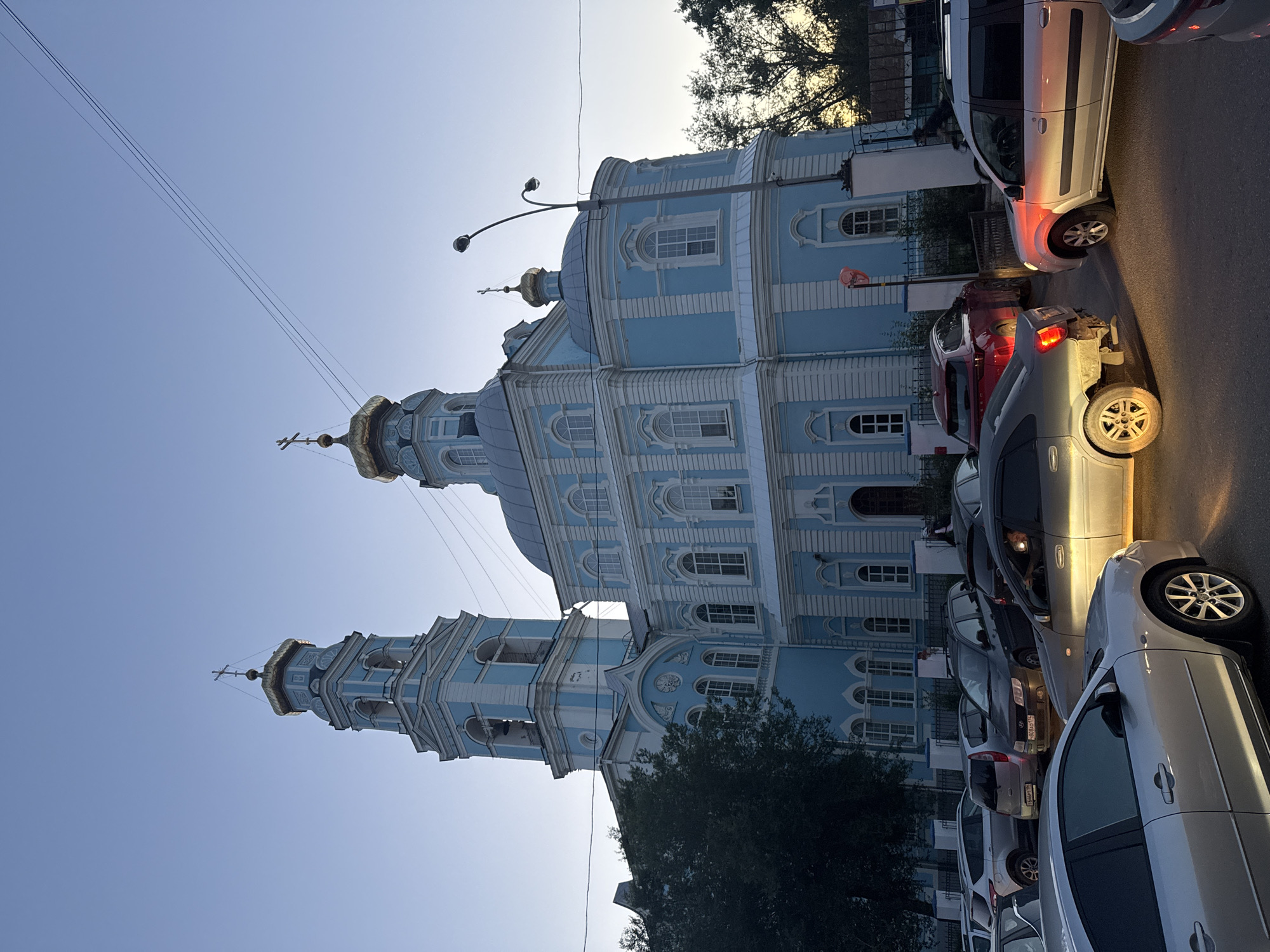 The image shows a light blue Orthodox church with golden domes and a bell tower. The church is situated in a city environment, with several cars parked and driving in front of it. The sky is clear and blue, suggesting it is either early morning or late afternoon. The overall scene appears to be a street in Yekaterinburg, Russia, possibly near a prominent religious site. There are no visible people clearly depicted or performing distinct actions, but the presence of cars suggests an active urban setting. The lighting indicates a time of day with soft, diffused sunlight.