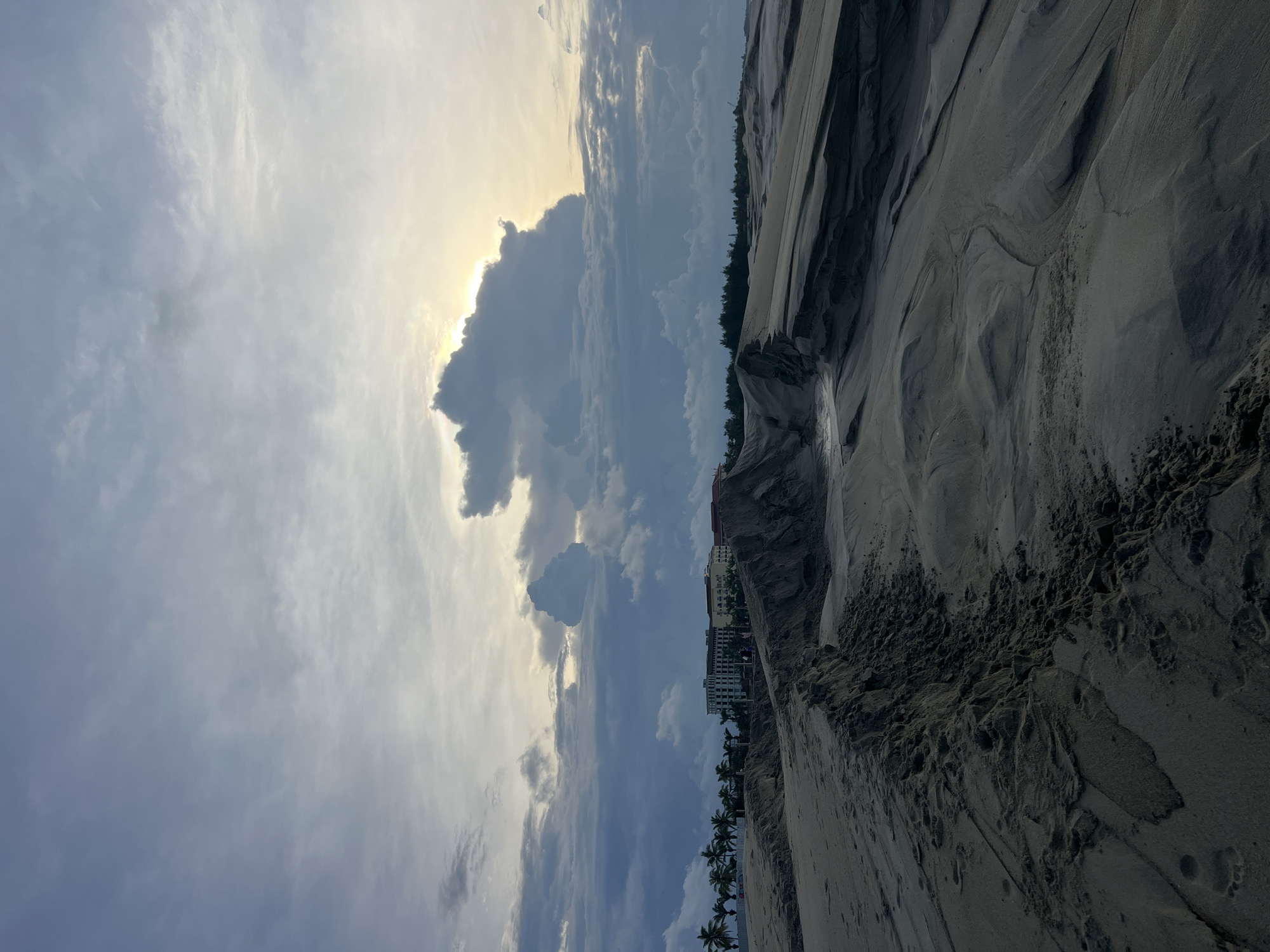 This media file depicts a scenic view of a sandy beach with a backdrop of dramatic clouds and a distant resort. The image is oriented vertically, suggesting a portrait mode capture or a rotated landscape.
The foreground is dominated by textured sand, showing patterns likely created by wind or water erosion. Several indentations, possibly footprints or small holes, are visible in the sand, adding a sense of the beach's natural landscape.
In the middle ground, a line of palm trees is faintly visible, hinting at a tropical or subtropical environment. Behind the trees, a multi-story building, appearing to be a hotel or resort complex, is situated on a cliff or elevated area overlooking the beach. The architecture suggests a modern design with multiple balconies.
The sky is a prominent feature, filled with a mix of light and dark clouds. The sun is partially obscured by a large, dark cloud formation, creating a dramatic contrast between light and shadow. The light filtering through suggests it is either early morning or late afternoon, during the golden hour. The weather appears to be partly cloudy, with a chance of rain or a recent downpour given the dark clouds.
The scene evokes a sense of tranquility and natural beauty, with the power of the sky contrasting with the stillness of the resort. The location is identified as Hoi An, Vietnam, which aligns with the presence of palm trees and coastal resorts. No visible text is present in the image.