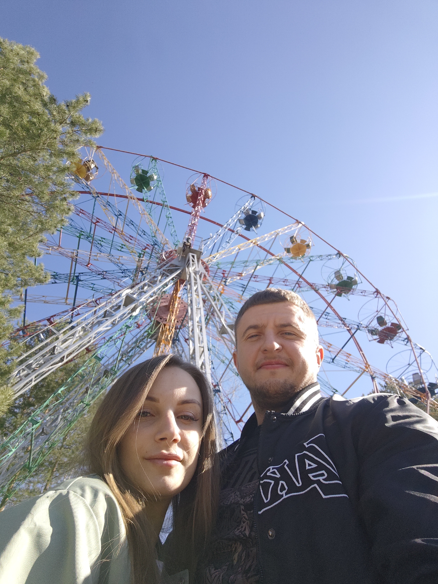 A selfie taken by a man and a woman in front of a Ferris wheel. The Ferris wheel has a white frame with colorful gondolas (red, yellow, green, and blue). A tree is on the left side. The sky is clear and blue.
The people are in the foreground. The woman has long brown hair. The man is wearing a black jacket with white lettering.
The location is likely Chaikovsky, Russia, based on the Ferris wheel.