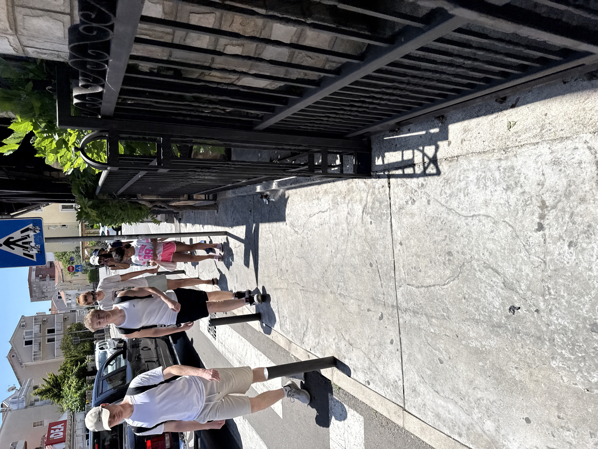 A group of five people, including three adults and two children, are walking on a sidewalk on a sunny day in Budva, Montenegro. The group is walking towards the camera, with two young males in the foreground. The first young male is wearing a white t-shirt, beige shorts, and a white baseball cap, and is carrying a black backpack. He is walking on the sidewalk. Behind him, another young male is wearing a white tank top, black shorts, and is carrying a black backpack. He is walking on the sidewalk. A woman with red hair, wearing a white t-shirt and light-colored shorts, is walking behind the two young males. A girl with dark hair wearing a pink t-shirt and pink shorts, holding a white bag, is walking in front of the woman. Another woman with dark hair and sunglasses is walking behind the girl. The sidewalk is made of concrete and has some cracks and stains. There are several black bollards along the sidewalk. To the left of the sidewalk, there is a black car and some buildings in the background, including a building with an "IDEA" sign. To the right of the sidewalk, there is a black metal fence and a stone wall. The sky is blue and clear, indicating good weather.